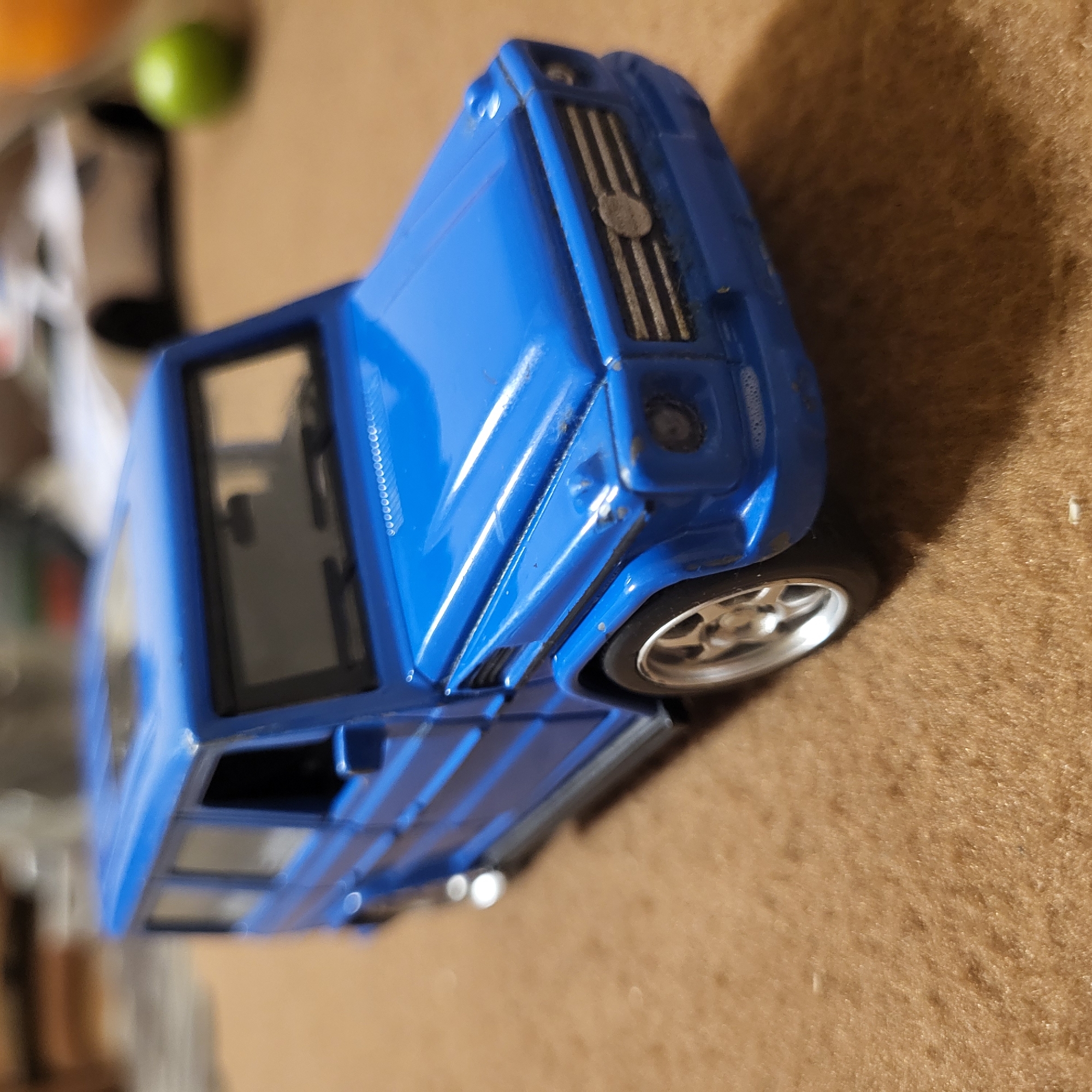 A blue toy car, possibly a model of a Mercedes-Benz G-Wagen, is positioned at an angle on a brown surface. The toy exhibits visible details such as the front grille, wheels, windows, and body lines. The background includes blurry objects, potentially a green lime. The location is not evident from this image and there is no indication it is in Yerevan, Armenia.