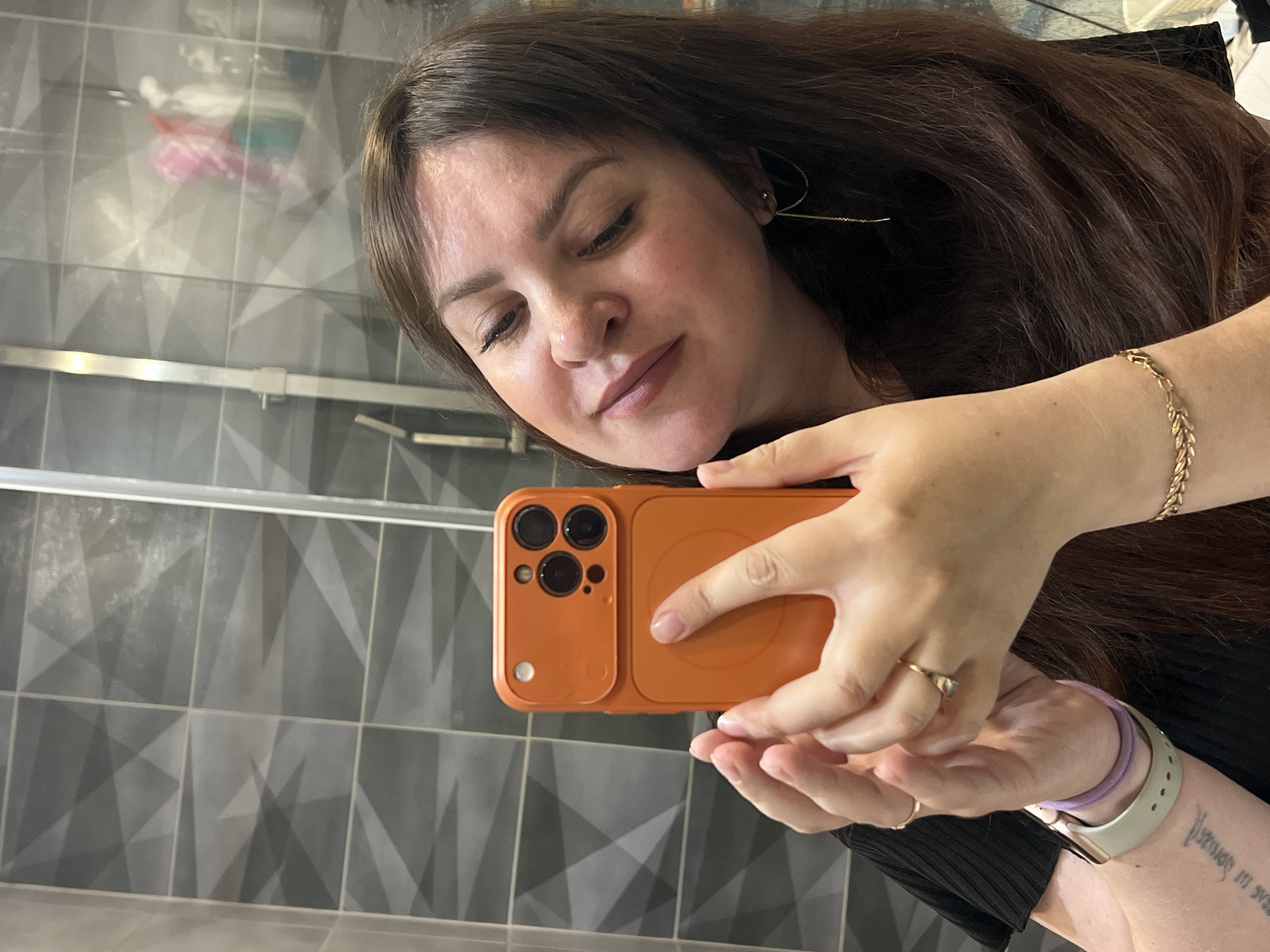 A woman takes a selfie in a bathroom with patterned grey and white tiles. The woman has brown hair and is wearing a gold bracelet, earrings, and rings. She is holding an orange iPhone with a MagSafe case. A watch with a lavender band is visible on her left wrist. The location is not discernible.