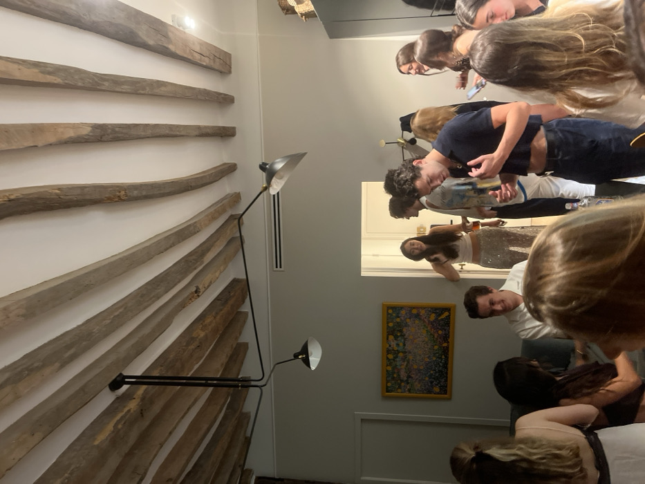 The image depicts an indoor social gathering of approximately 10-12 young adults in what appears to be a residential space, characteristic of an apartment in Paris, France. The ceiling features prominent, curved, exposed wooden beams against a white surface, from which a black, multi-arm ceiling lamp with two visible cone-shaped shades is suspended. A horizontal air vent is visible high on the right wall. A framed abstract painting with multicolored splatters on a dark background hangs on the left wall.
In the mid-ground, a male with dark, curly hair stands centrally, wearing a dark blue cropped polo shirt and dark trousers, looking down at his hands. Behind him, another male with dark, curly hair stands in a light-colored patterned shirt and white bottoms. To the left, a male in a white collared shirt is seated, smiling. In a doorway towards the rear-left, a female with long dark hair stands, wearing a white crop top and silver sequined pants or a long skirt, holding a beverage in a glass and smiling. Several other individuals are present in various states of interaction and posture. A person with blonde hair wearing a light-colored camisole with black lace is visible from the back in the far left foreground. Another person with blonde hair is seen from behind in the central foreground, with a clear bottle visible on a surface in front of them. A group of females are visible on the right side of the frame, some appearing to hold drinks and engage in conversation. The scene suggests a casual social event with multiple individuals standing, sitting, and interacting.