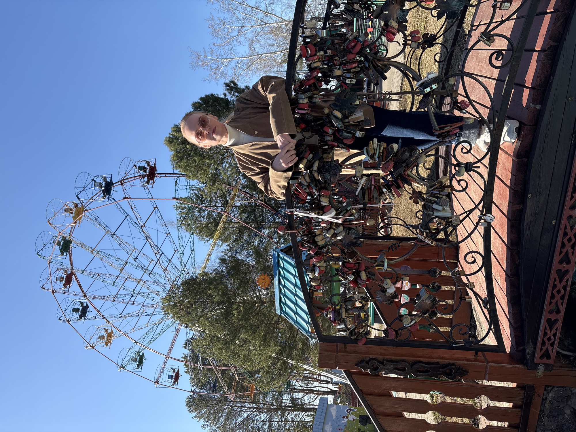 An outdoor, vertically oriented full-length shot features a person posing beside a metal railing adorned with numerous 'love locks,' with a large Ferris wheel in the background against a clear blue sky. The person, positioned in the middle ground, has light-colored hair pulled back, wears round sunglasses, a light-colored high-neck top, and a brown jacket. They lean on the railing, looking towards the camera with a subtle smile, their hands clasped.
The metal railing, likely part of a bridge or scenic overlook in Chaikovsky, Russia, is densely covered with a multitude of padlocks of various colors and designs, including red, black, gold, and heart-shaped locks, symbolizing love and commitment. Behind the person and the railing, a large, colorful Ferris wheel dominates the left side of the frame, its cabins in shades of red, yellow, blue, and green.
The background also includes several evergreen trees and some bare-branched deciduous trees, suggesting late winter or early spring. A small structure with a green corrugated roof and a yellow sun decoration is visible behind the love-lock-covered railing. The bright sunlight and deep blue sky indicate a clear, sunny day. The scene captures a moment of leisure in what appears to be a park or amusement area.