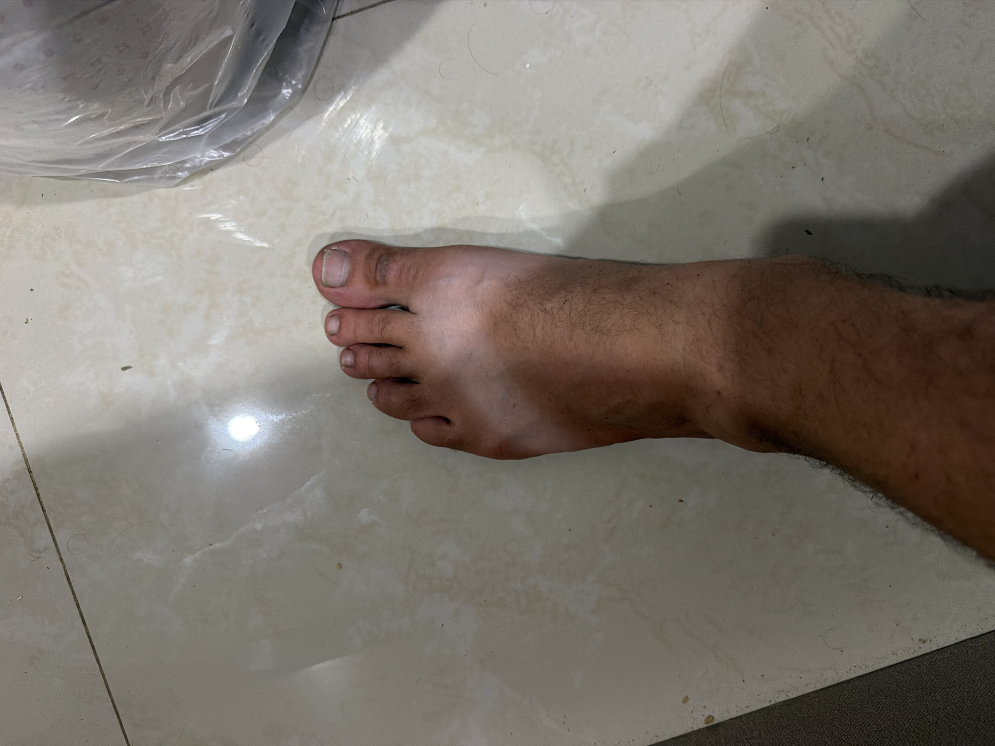 A left foot with a distinct tan line from being exposed to the sun, with the top of the foot being lighter than the rest, is visible. The foot is positioned on a light-colored tiled floor. The environment appears to be indoors, with a bright light source reflecting off the tiles. There is a portion of what appears to be a plastic bag in the upper right quadrant of the image. The image was taken in Baan Mae Nam, Thailand.
