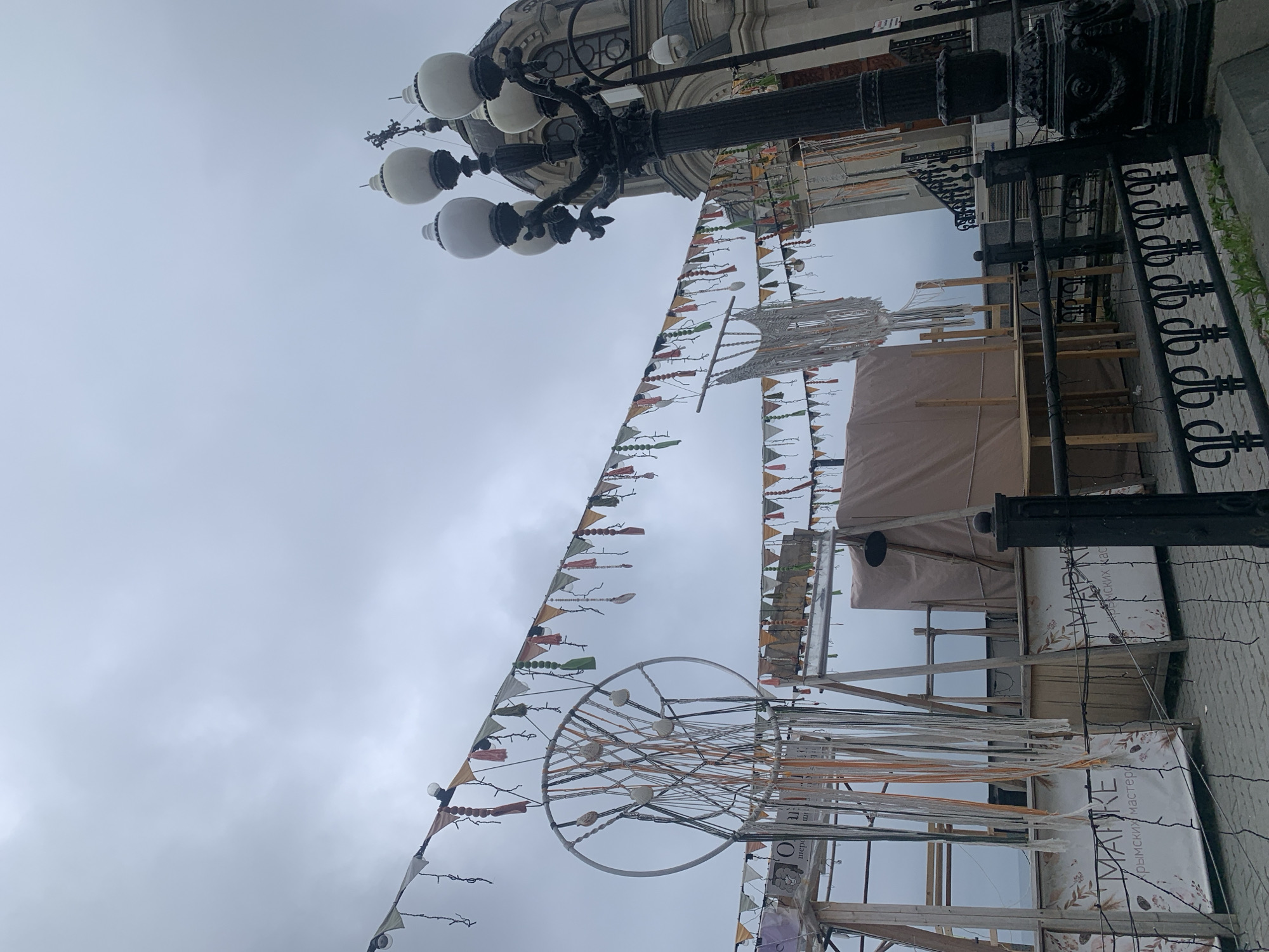 The image depicts a street scene with decorations and a street lamp under an overcast sky. Banners with small triangular flags are strung across the street, and a metal structure with a wheel-like element and hanging ribbons is present. The street lamp is a multi-globe style with ornate detailing. A building with architectural details is partially visible in the background, and a wrought-iron fence runs along the sidewalk. A temporary structure, possibly a market stall, is set up in the street. The language on the storefront sign is not English. The location is specified as the city of Unknown, Unknown.