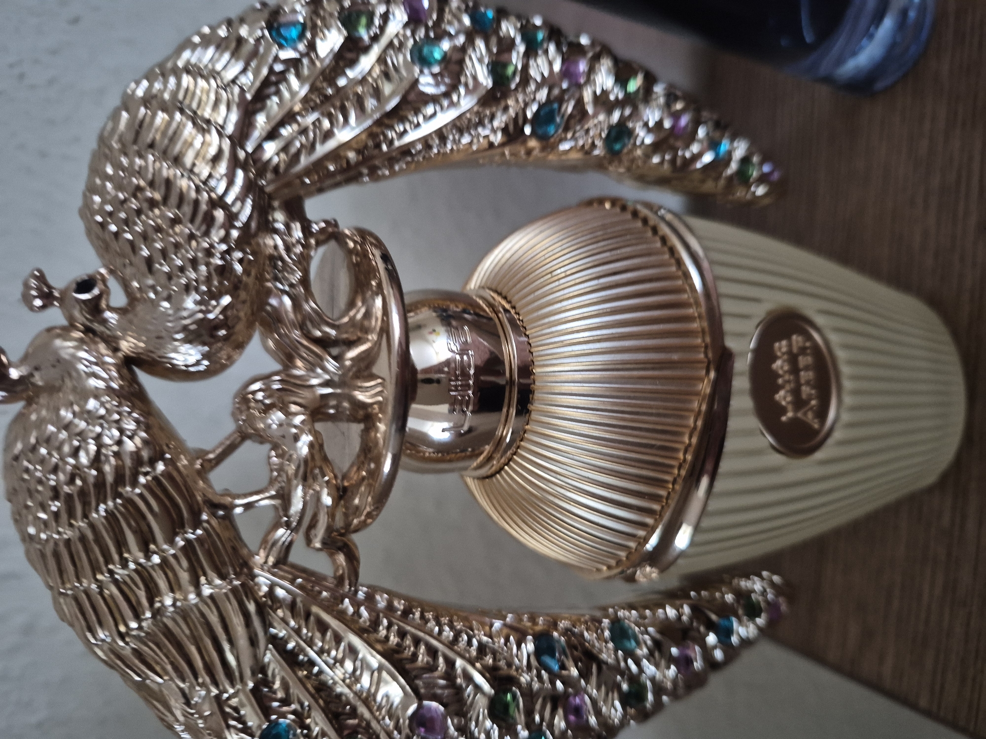 The image shows a close-up, eye-level, indoor shot of a decorative perfume bottle with two gold peacocks embracing the top. The bottle is a light beige color with vertical ridges and a gold, ridged cap. The peacocks are intricately detailed, with feathered wings adorned with colorful gemstones, including blue, green, and purple. The background is a neutral, light gray. The perfume bottle is placed on a wooden surface, and a dark blue bottle is partially visible to the right. The lighting is bright, casting some reflections on the metallic surfaces. The scene is static, with no people or animals present.