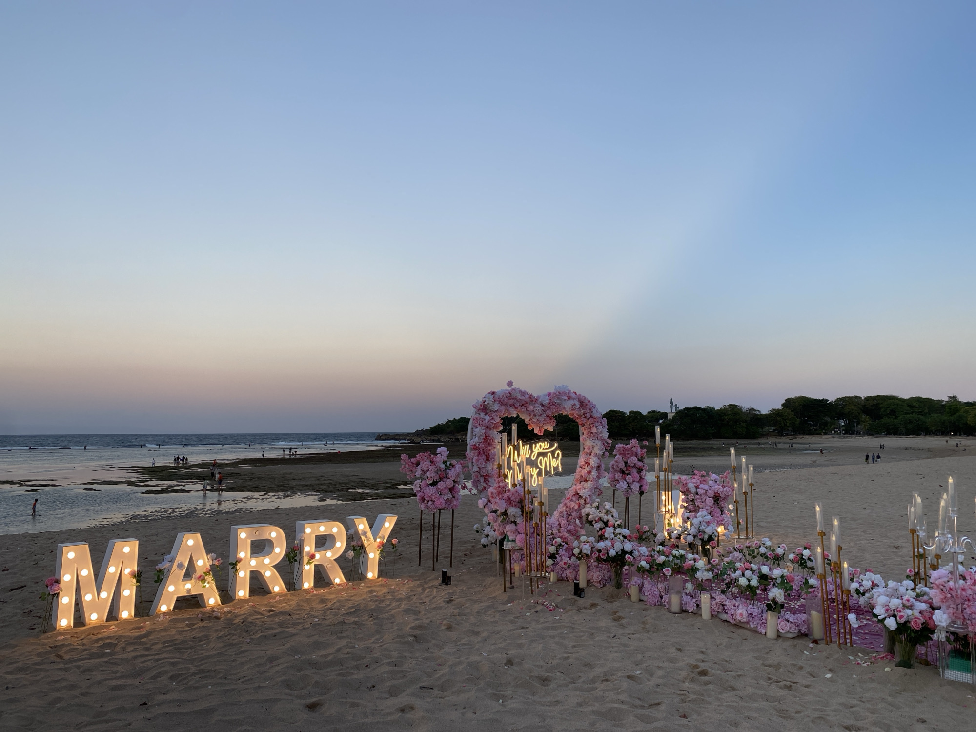 This is a wide, low-angle shot of a beach proposal setup at sunset in Kuta Selatan, Indonesia. The sand is a light brown, and the ocean is visible in the background, with gentle waves lapping at the shore. The sky is a gradient of soft blues and pinks, with light rays emanating from the horizon.
In the foreground, large, illuminated marquee letters spell out "MARRY". Each letter is white and outlined with a string of warm-toned lights. Pink and white faux flowers are artfully placed around the base of these letters.
To the right of the "MARRY" sign is a large heart-shaped archway, also adorned with a profusion of pink and white flowers. Inside the heart, a neon sign glows with the words "Will you marry me?". The arch is flanked by tall, gold-colored stands holding bouquets of pink and white flowers and clear glass candle holders with lit candles. Further to the right, a decorative arrangement of pink and white flowers in vases and clear cylinders extends along the sand.
In the middle ground, several people can be seen walking along the beach and in the shallow water. The water is calm, with sandbars visible. In the far background, a tree line separates the beach from what appears to be a town or resort area.
