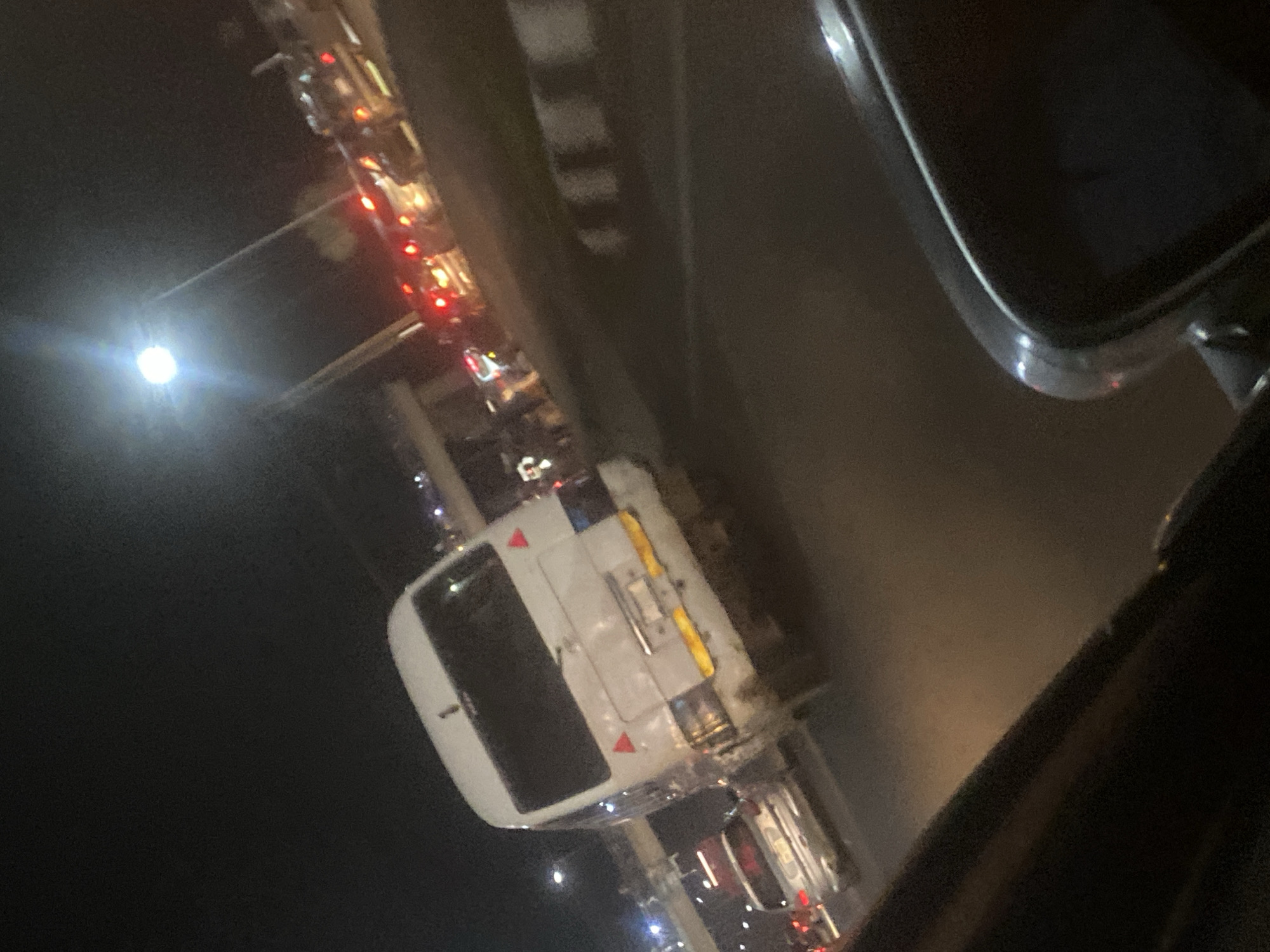 At night in Gosa, Nigeria, the scene, viewed from inside a vehicle, shows a traffic jam with numerous vehicles, including a white van with reflectors, on a road illuminated by streetlights. Red taillights are visible on many of the cars. The environment is dark and urban.
