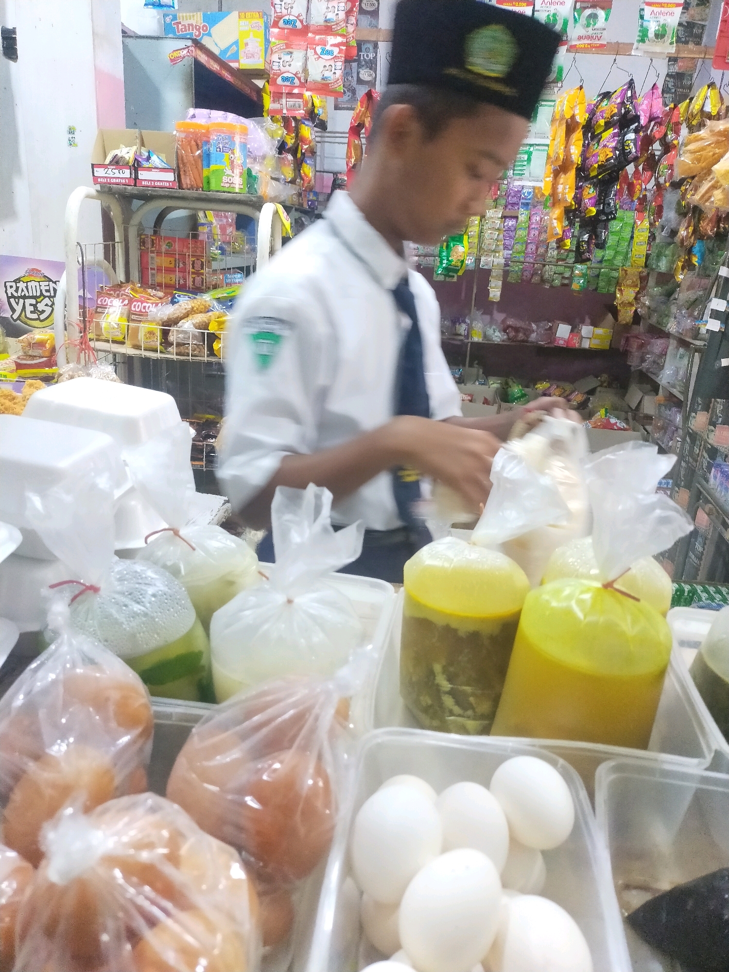 In the city of Mojodanu, Indonesia, a young boy wearing a white shirt, dark blue tie, and a black peci is seen working at a small convenience store. The store is filled with shelves stocked with various snacks and drinks, including packaged goods like Tango wafers, colorful candy bags, and ramen. In the foreground, several food items are displayed in clear plastic bags and containers. There are bags containing what appear to be hard-boiled eggs, and containers with liquid food items, some of which are yellow and green in color. The boy is in the process of handling food items, possibly packaging or arranging them. The store has a cluttered yet organized appearance, typical of a small, local establishment.