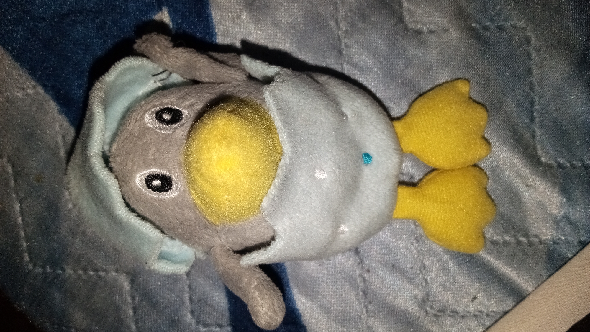 A close-up, overhead shot shows a gray plush toy penguin wearing a light blue outfit with white polka dots. The penguin has large, black and white eyes and a bright yellow beak. Its yellow webbed feet are visible at the bottom of the frame. The toy is lying on a blue and white patterned fabric, possibly a blanket or bedspread.