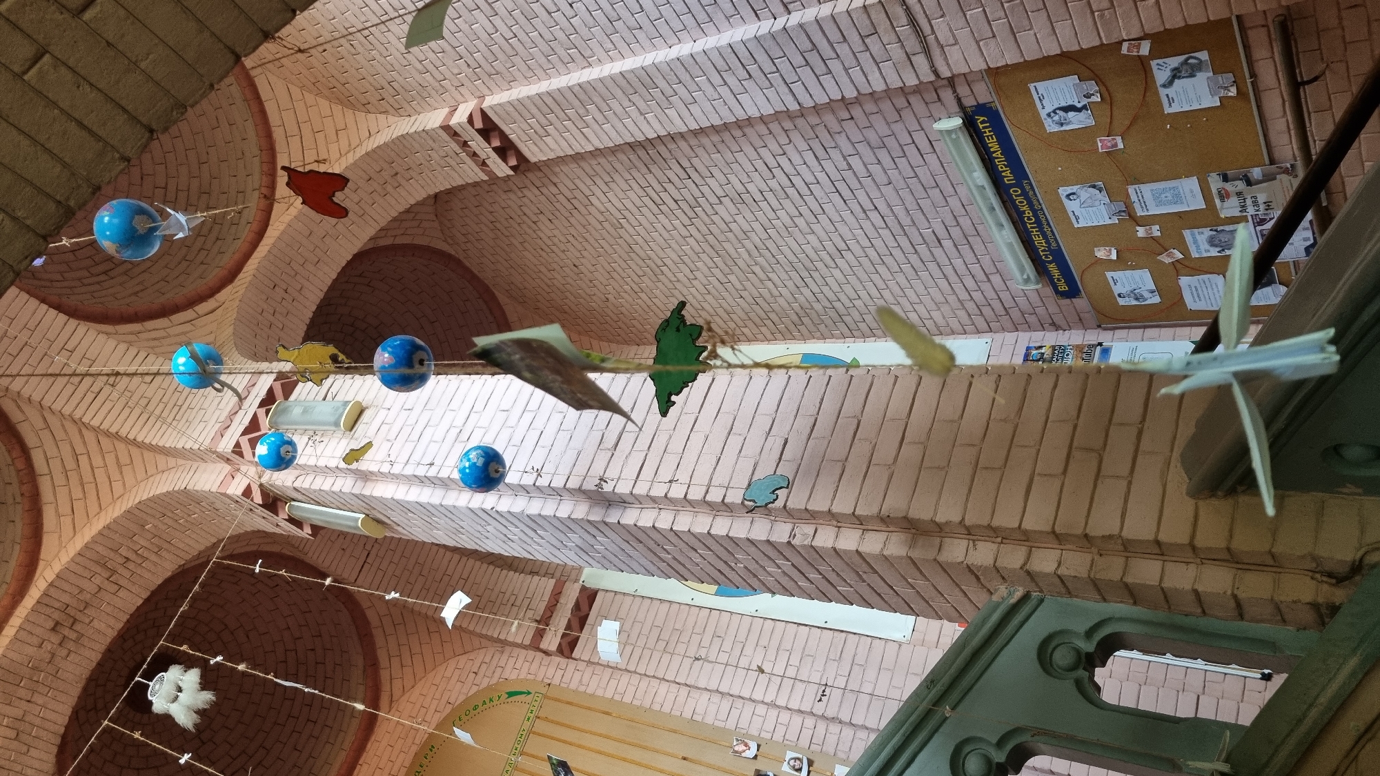 The image shows an interior scene, likely a school or university hallway, with a prominent brick wall and arched ceiling. Several strings are hanging from the ceiling, adorned with various objects including blue spheres resembling globes, paper cutouts of animal shapes and continents, folded paper airplanes, and what appear to be small photographs. A bulletin board is mounted on the brick wall, displaying posters and papers. In the foreground, a dark green wooden railing with ornate carvings is visible. The lighting suggests an indoor environment with artificial light sources, possibly fluorescent tubes visible on the ceiling. The architecture, specifically the brickwork and arches, suggests a building with some age and character. The text on the bulletin board and posters is in Ukrainian. The bulletin board has a title that reads "ВІСНИК СТУДЕНТСЬКОГО ПАРЛАМЕНТУ" (Herald of the Student Parliament), with a subtitle "Географічного факультету" (of the Faculty of Geography). This strongly indicates the location is within the Geography Faculty of a university in Chernivtsi, Ukraine, specifically at Chernivtsi National University named after Yurii Fedkovych.