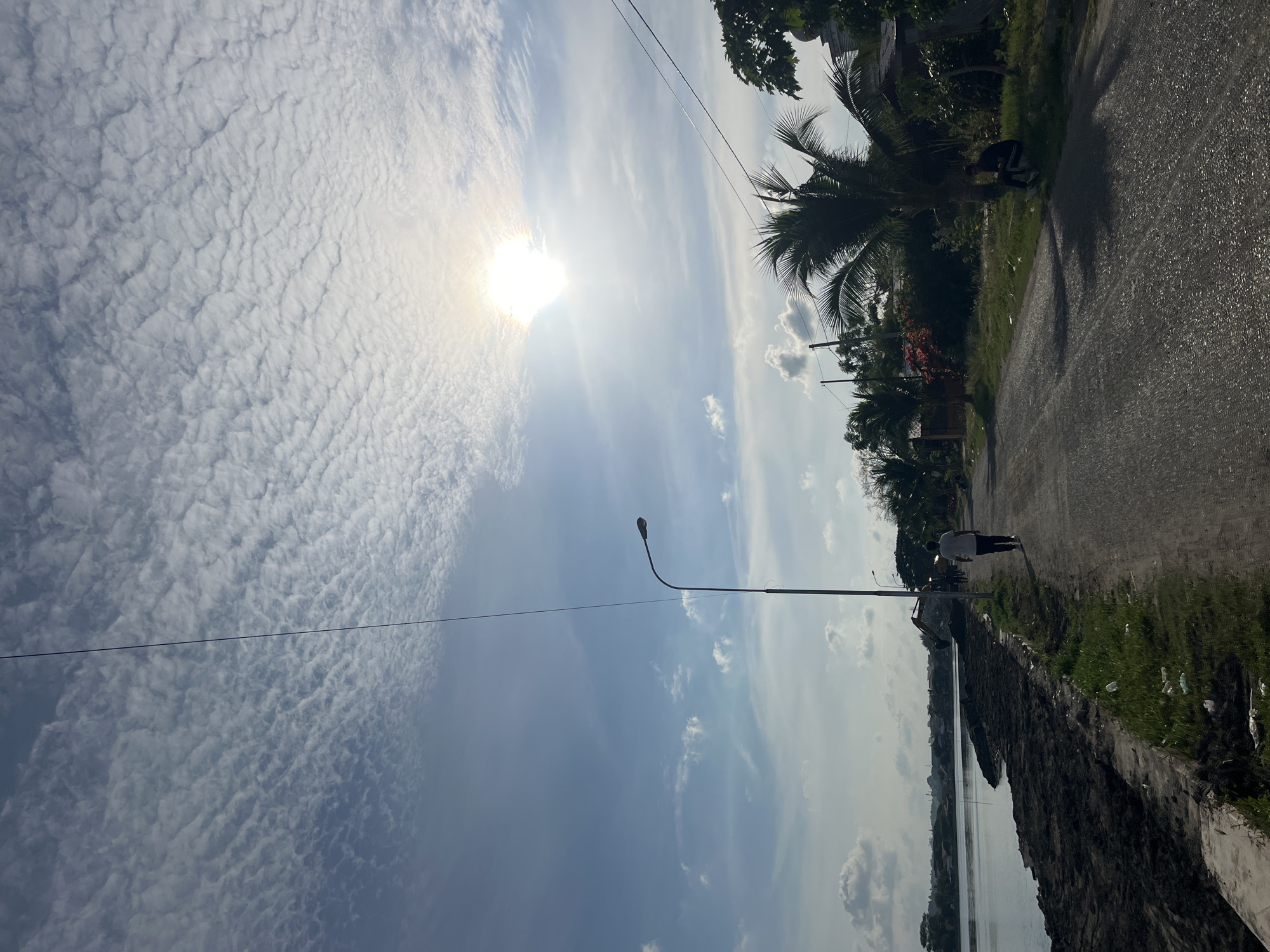 The photograph depicts a scene in Lhokseumawe, Indonesia. A street, running vertically, is bordered by a body of water on the left and vegetation on the right. A person is walking along the street. A street lamp is visible between the water and the road. The sky is partially covered with altocumulus clouds and the sun is partially obscured by the clouds.