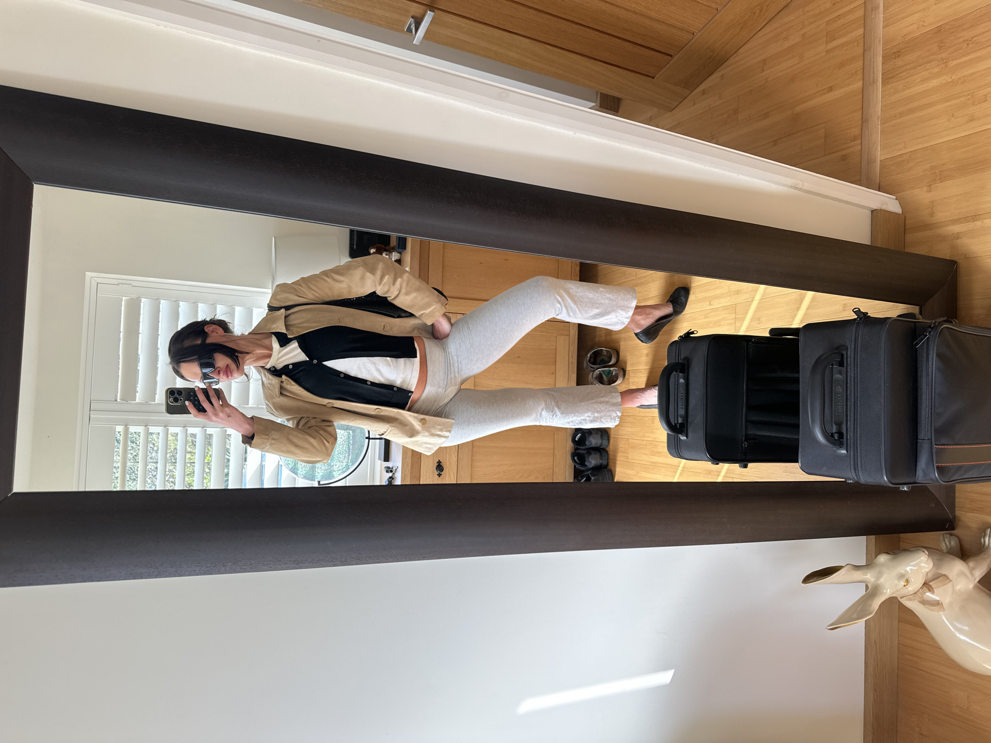 A full-length mirror selfie captures a woman posing indoors in a location within Wealden, United Kingdom. The woman, with dark hair pulled back and wearing dark rectangular sunglasses, stands facing the mirror. She is dressed in a light-colored cropped top, a black button-up cardigan, and a beige casual jacket. Her lower attire consists of light grey, flared-hem trousers and black ballet flats. She holds a dark smartphone, visible with its camera lenses, in her raised right hand to take the photo, while her left hand rests on her hip. A tattoo is visible on her right wrist.
In front of the mirror, two black soft-shell suitcases are stacked. To the left of the luggage, on the light wooden floorboards, is a large, glossy, beige ceramic rabbit statue featuring golden ears and a small bow around its neck. Behind the woman's reflection, a light wood dresser or cabinet with dark handles is visible, topped with small items. Above this, a window with white plantation shutters is reflected, revealing green foliage outside. Several pairs of shoes, including dark athletic shoes, are lined up on the floor beneath the dresser. The room has white walls, and a wooden door with a silver handle is partially reflected on the far right. Natural light illuminates the scene, casting shadows on the wall to the left of the mirror.