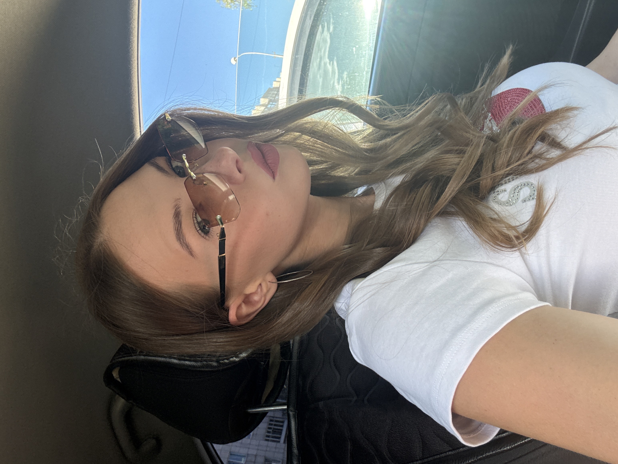 A young Caucasian female with long, wavy brown hair and fair skin is seated in the driver's seat of a car, looking directly at the camera. She is wearing a white t-shirt with a pink heart design embellished with rhinestones and rimless sunglasses with tinted lenses. A silver hoop earring is visible in her left ear. The car's interior is visible, including the black leather seat and headrest. The background shows a sunny sky and buildings, suggesting an urban setting, potentially Perm, Russia, given the presence of brick apartment buildings characteristic of the region. The image is taken from a slightly low angle, with sunlight glinting off the car window.