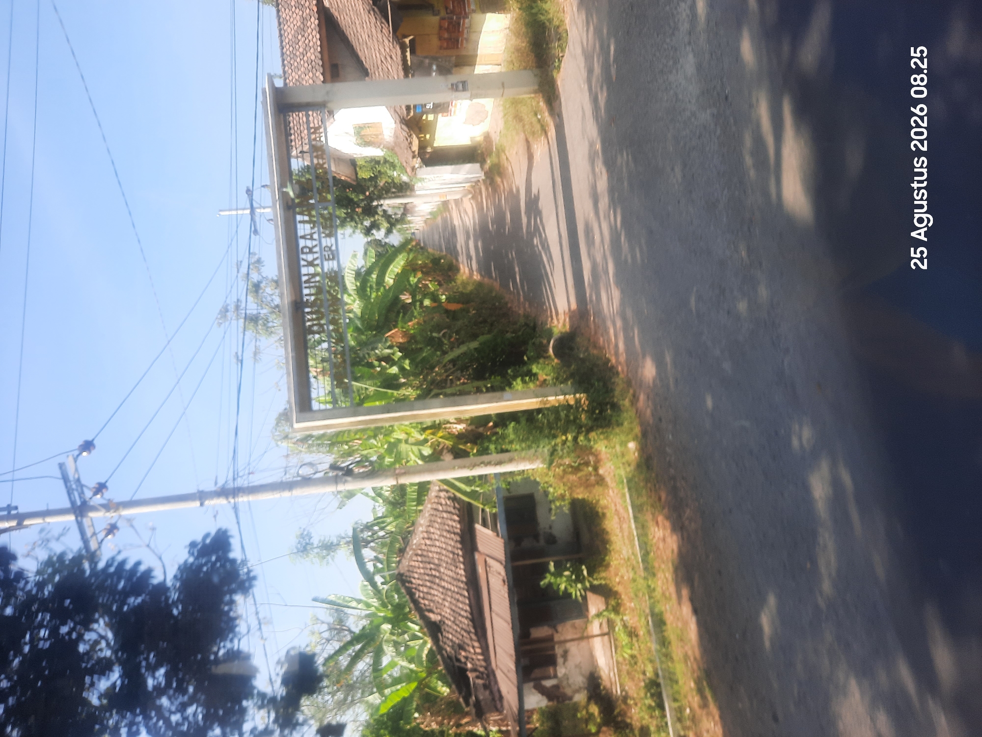 The image captures a rural scene in Sukokerto, Indonesia, with a road leading through lush vegetation. A prominent overhead archway is visible, bearing the inscription "DUSUN KRAJAN". To the left of the road, a dilapidated house with a broken tiled roof stands partially obscured by banana trees. Further down the road, several houses with tile roofs are visible on the right side. The sky is clear and blue. The image is dated "25 Agustus 2026 08.25".