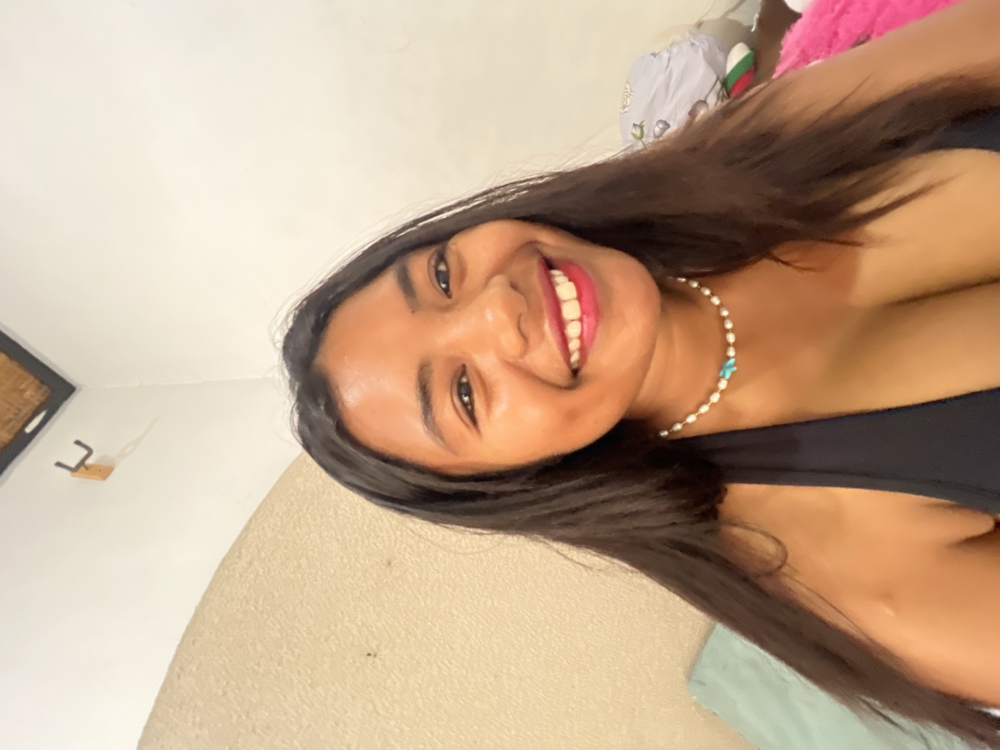 The image shows a close-up of a young woman with dark hair, smiling and looking directly at the camera. She is wearing a black halter top and a pearl necklace with a blue pendant. The background is a textured wall, and a glimpse of a bed with pillows and blankets is visible on the left. A hook is mounted on the wall in the upper left corner. The lighting suggests an indoor setting, likely in Denpasar, Indonesia, given the context.