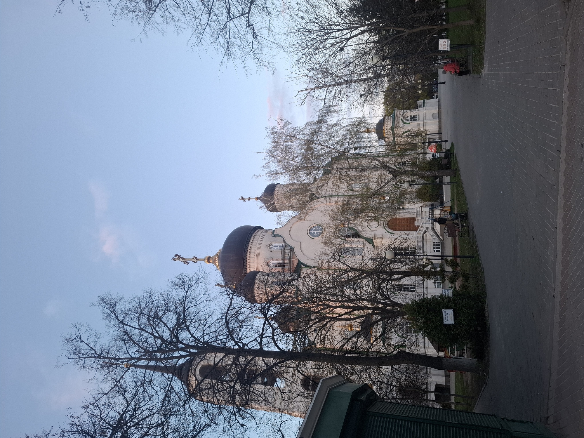 This image captures a serene outdoor scene featuring a prominent Eastern Orthodox church or cathedral complex, likely situated within a park. The main building, centrally located, is a large, white-plastered structure adorned with multiple dark, onion-shaped domes topped with golden Orthodox crosses. Its facade features decorative elements, arched windows, and distinct circular windows. To the far left, a tall bell tower or spire with another cross is partially visible, while another smaller domed building stands further back on the right.
The setting is characterized by numerous bare deciduous trees, their intricate branches framing the sky and parts of the architecture, suggesting late autumn or winter. Some evergreen shrubs are also visible among the trees. Paved pathways, one made of grey blocks and another of reddish-brown bricks, lead through the park.
A few people are present in the scene; one person in a bright red jacket is noticeable in the mid-ground on the main paved path, and another appears to be seated on a bench further down. The soft, diffused lighting and the pastel blue sky with faint streaks of pinkish clouds hint at either dawn or dusk, creating a tranquil atmosphere. A white sign with dark Cyrillic text, partially obscured by foliage, is visible next to the path, bearing the words "КОРМЛЕНИЕ ГОЛУБЕЙ" (Feeding of pigeons), indicating a common park regulation. The overall impression is one of peaceful contemplation in a historic religious site.