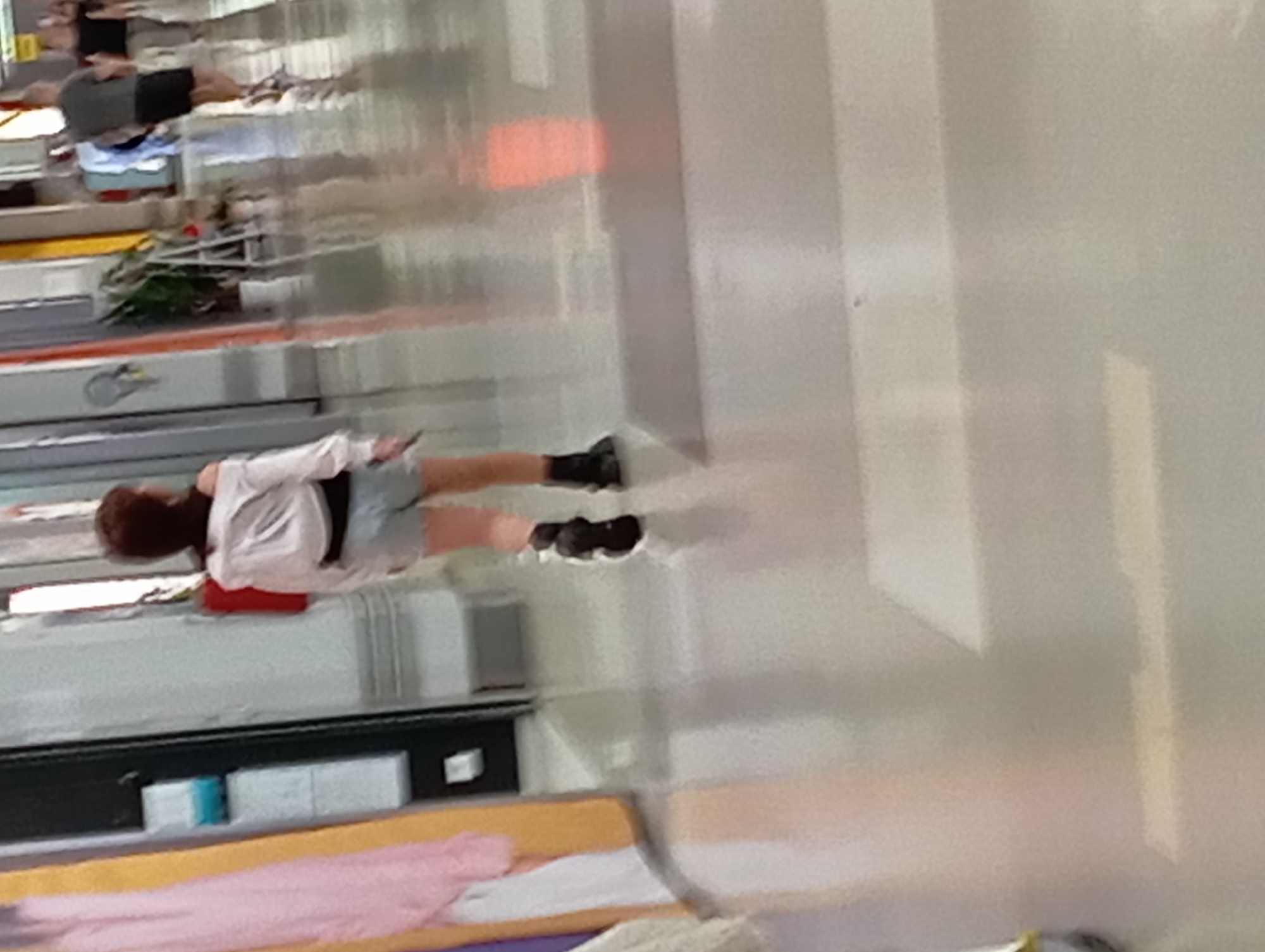 The image shows the interior of a public space, possibly a shopping mall or a walkway in a city. In the foreground, a person with short brown hair is walking away from the camera. They are wearing a white off-the-shoulder shirt, light-colored shorts, and dark boots. To the left of the person, a display of clothing, primarily pink and yellow, is visible. In the background, other people are walking in the opposite direction. One individual wears a dark t-shirt and shorts, while another wears a light blue shirt. The environment appears to be well-lit with a polished floor that reflects the light. There are no distinct architectural features or signage that would indicate a specific location within Nitra, Slovakia. The focus is on the person in the foreground and their attire.