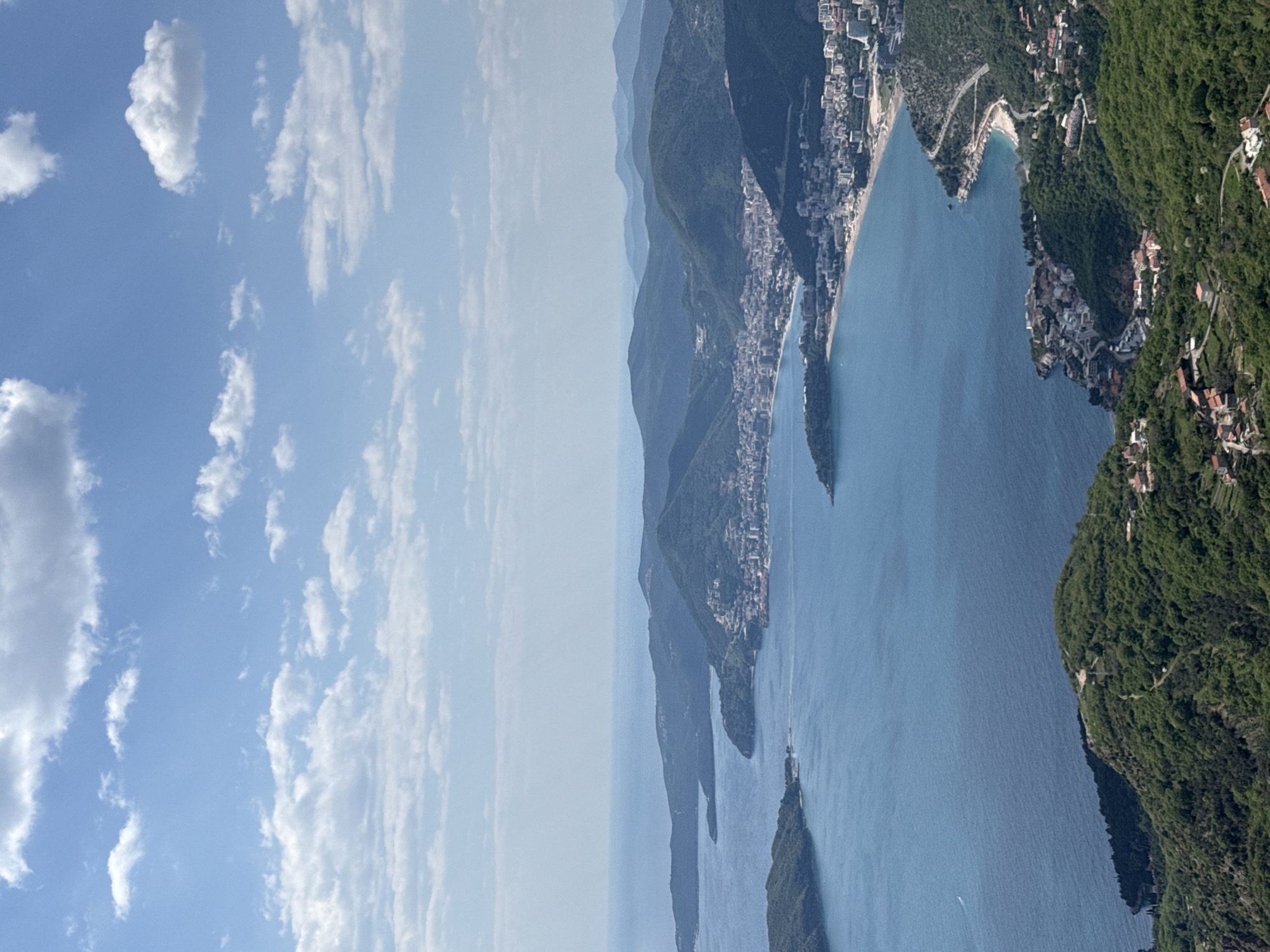 The image depicts a coastal landscape with a blend of natural and urban elements. On the right side, mountains covered in lush green vegetation rise sharply, interspersed with residential buildings and roads. The terrain slopes down to meet the blue water of the sea, which stretches out to the center of the frame. Several small islands are visible in the water, with buildings clustered along the coastline. The sky occupies the left side of the image, filled with scattered white clouds against a clear blue backdrop. The scene suggests a coastal city or town nestled among mountains, typical of Mediterranean or Adriatic regions. Without more context, the location in the city of Unknown, Unknown cannot be identified definitively.