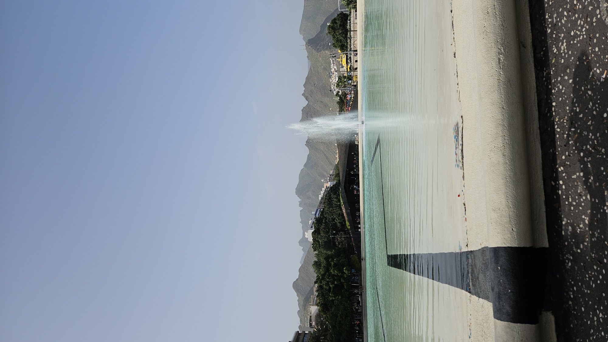 Here is a description of the image:
This photograph, taken in Santa Cruz de Tenerife, Spain, showcases a large, rectangular water feature with a prominent central fountain spraying water high into the air. The water has a slightly greenish tint. The fountain is situated against a backdrop of layered mountains, dotted with sparse vegetation and buildings that transition into more dense trees near the base. The sky is a pale blue, suggesting clear weather. The foreground includes elements of the water feature’s edge and surrounding pavement. The edge appears to be painted white with a wide black stripe along the lower section. The pavement has a dark, speckled texture and is in the immediate foreground.