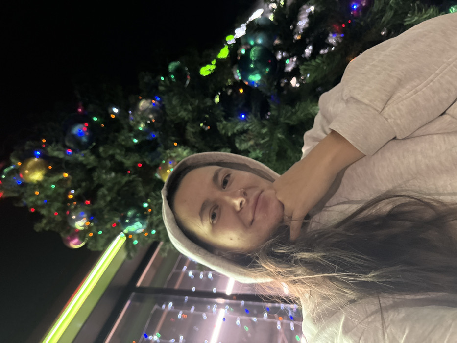 A woman poses in front of a Christmas tree. She wears a gray hoodie and rests her chin on her hand. Long brown hair falls across her shoulder. The Christmas tree is decorated with colorful lights and ornaments, with a window reflecting the lights. A yellow glow illuminates the left side of the image, contrasting the dark background. The picture is taken at a slight angle, creating a sense of casualness.