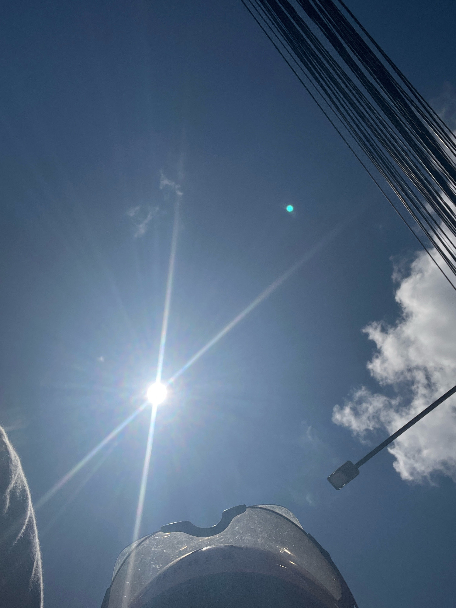 The image depicts a low-angle, upward view of a clear blue sky with scattered white clouds. The sun is prominently visible on the left side, creating a strong lens flare with bright rays extending outwards. A faint, circular green light artifact is present in the upper middle portion of the sky. In the foreground, the top edge of a motorcycle helmet is visible, along with a portion of a person's shoulder on the far left. A utility pole with several wires runs diagonally across the upper right of the frame, and a street light is attached to a pole extending from the right side of the image. This scene is consistent with an outdoor environment, possibly a street or roadside, in Denpasar, Indonesia, where such infrastructure and clear skies are common.