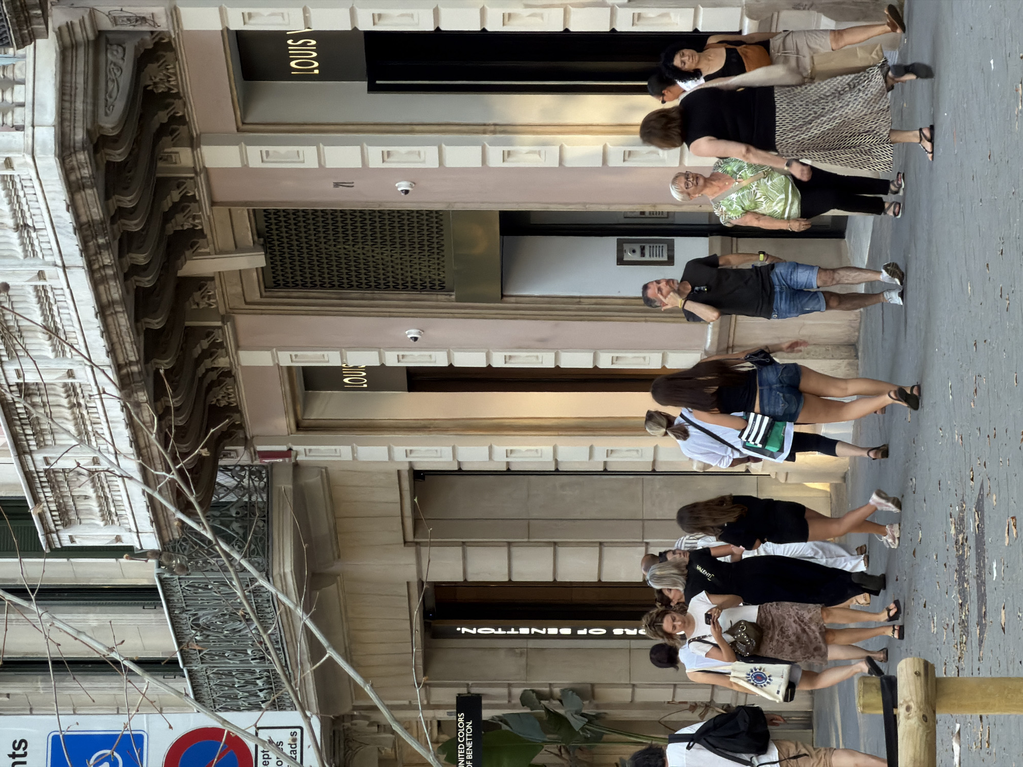 The image depicts a street scene in Barcelona, Spain, with several pedestrians walking on a sidewalk in front of shops. The architecture features ornate balconies and decorative stone detailing on the building facades. The shops visible include Louis Vuitton and United Colors of Benetton. Several individuals are captured in motion, engaged in various activities such as walking, looking at their phones, or gesturing. The ground is paved, and fallen leaves are scattered across the sidewalk. The lighting suggests it is daytime, possibly late afternoon.