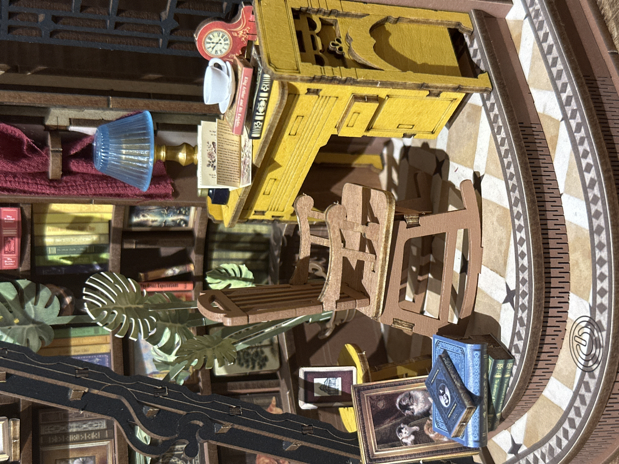The media file displays a meticulously crafted miniature diorama or book nook, featuring an indoor setting that resembles a cozy, old-world study or library. The scene is viewed from an elevated perspective, slightly tilted.
Central to the composition is a bright yellow wooden desk, intricately detailed with drawers and carvings. Perched on the desk are miniature accessories: a red and gold wall clock (displaying a decorative time), a small white teacup, several tiny books, and a blue fluted table lamp with a golden base. Adjacent to the desk, a wooden rocking chair and another chair are visible, suggesting a reading or working space.
The background and left side are dominated by bookshelves densely packed with miniature books of various colors, including red, green, yellow, and blue spines. Among the books and furniture, several green cut-out monstera leaves add a touch of lush greenery. A section of red textured fabric is draped on the left, hinting at a sofa or cushioned area.
In the lower right corner, a framed picture depicts two cats or kittens, and another smaller framed image is present. The floor features a distinct pattern of light and dark geometric shapes, including stars, and a curved border with ruler-like markings and a swirling design. A dark, ornate wooden structure, possibly a balustrade or decorative railing, occupies the foreground.
No people are visible in the scene. The overall impression is one of quiet contemplation and comfort, typical of a miniature library or study. The lighting appears artificial, highlighting the intricate details of the diorama.
Visible text includes "King Arthur and his Knights" on the spine of a red book, "The Great Expectations" and "Treasures of the Sea" on green book spines, and illegible text on open miniature books and a blue book in the foreground, some with small floral illustrations. The scene provides no specific visual cues for the user-specified location of Bobov Dol, Bulgaria, as it represents a generic, stylized miniature interior.