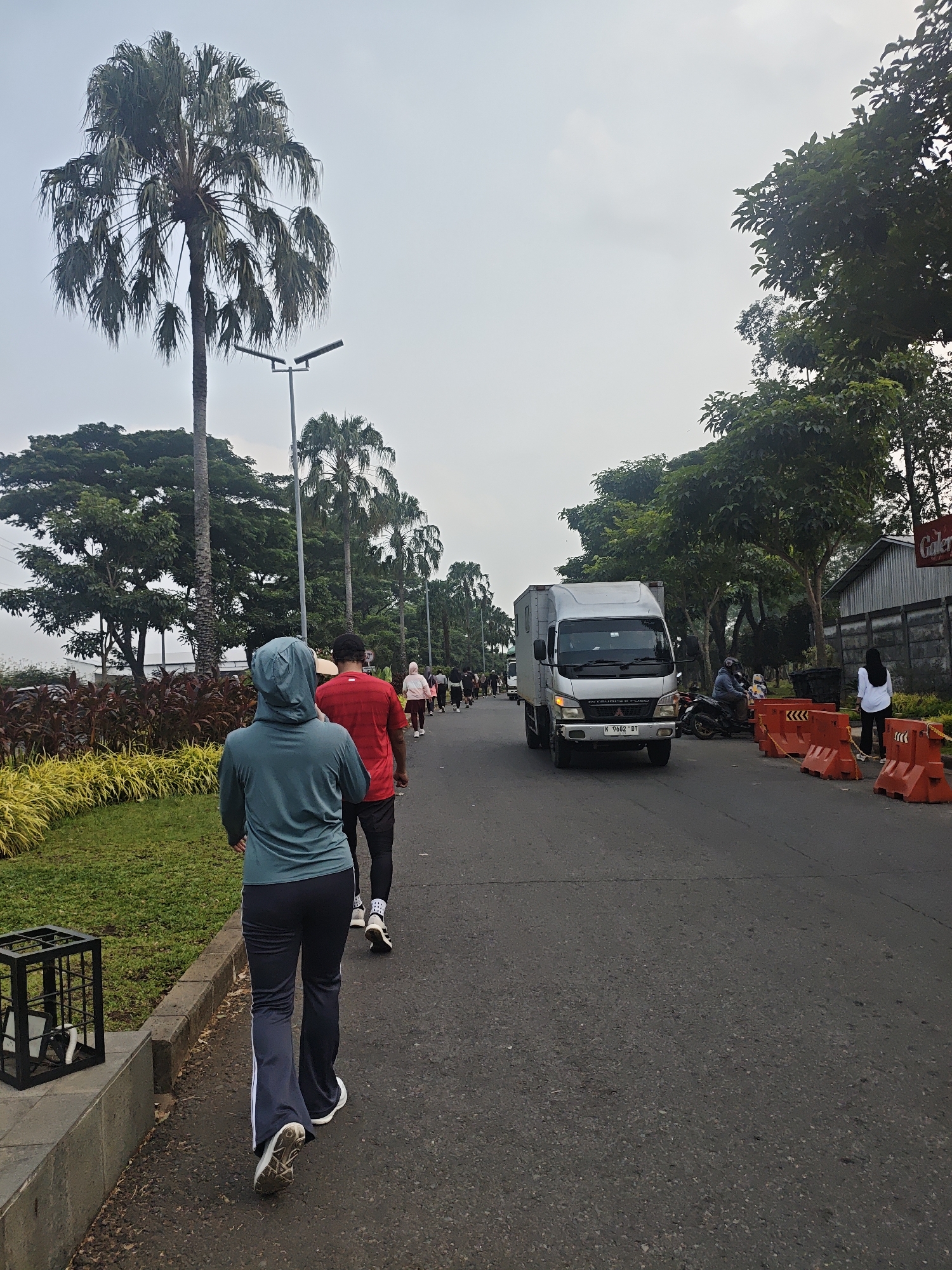 In Bacin, Indonesia, a group of people are engaged in an outdoor activity, likely a walk or run, along a paved road bordered by a grassy area with yellow and red foliage. In the foreground, a person wearing a teal hooded jacket and dark pants walks away from the camera. Behind them, another individual in a red shirt and dark pants is also walking. A silver box truck is proceeding down the road, followed by a line of people walking in the same direction. Orange traffic barriers are placed along the right side of the road, with a fence and trees in the background. The sky is overcast, and several tall palm trees are visible.