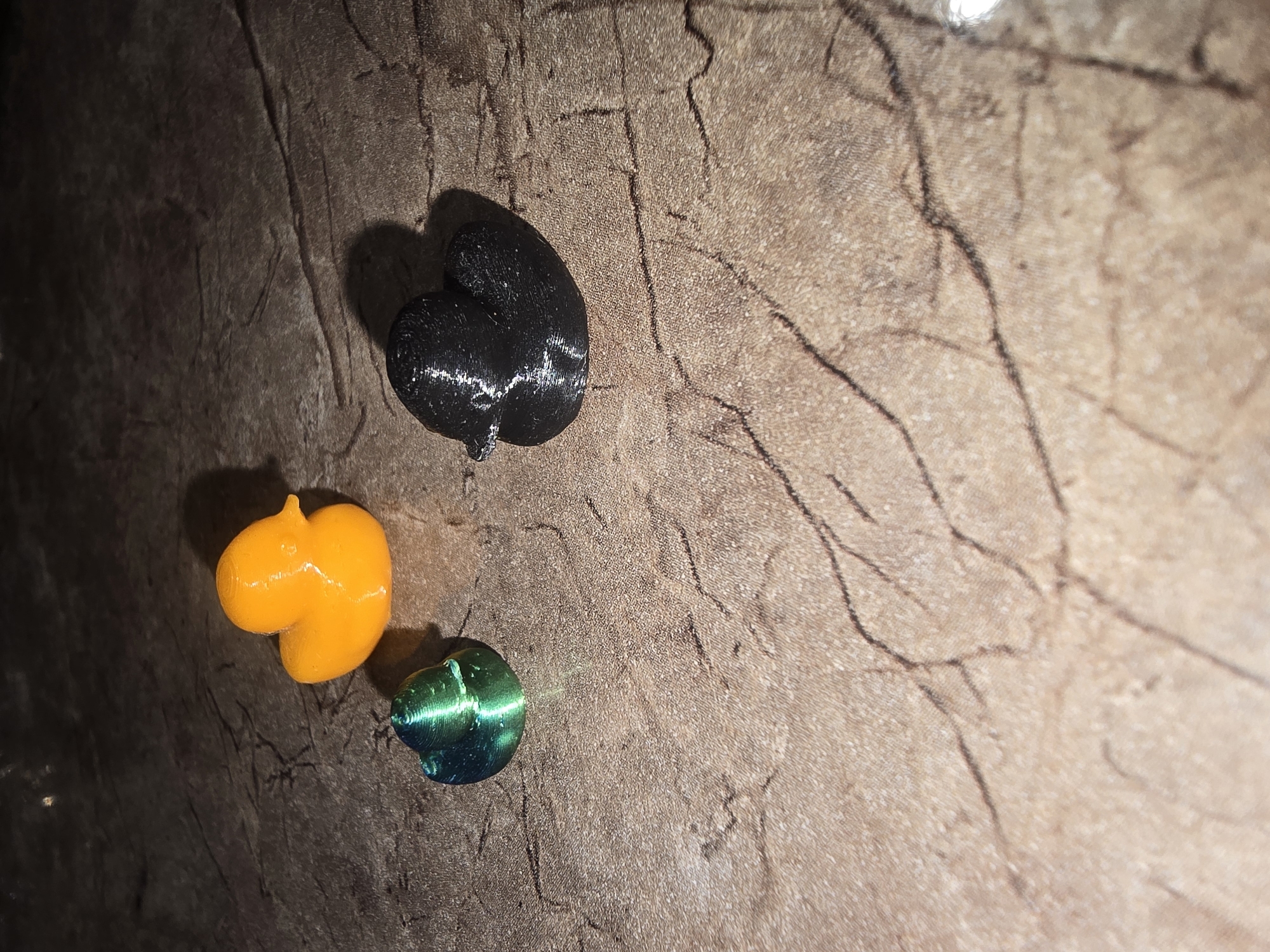 Three small figurines resembling ducks are arranged on a textured, dark brown surface with a marble-like pattern. From left to right, the figurines are: a small, metallic green duck; a medium-sized, bright orange duck; and a medium-sized, matte black duck. The lighting suggests the figurines are casting shadows. The location of this scene within Ulan-Ude, Russia, cannot be determined from the provided image.