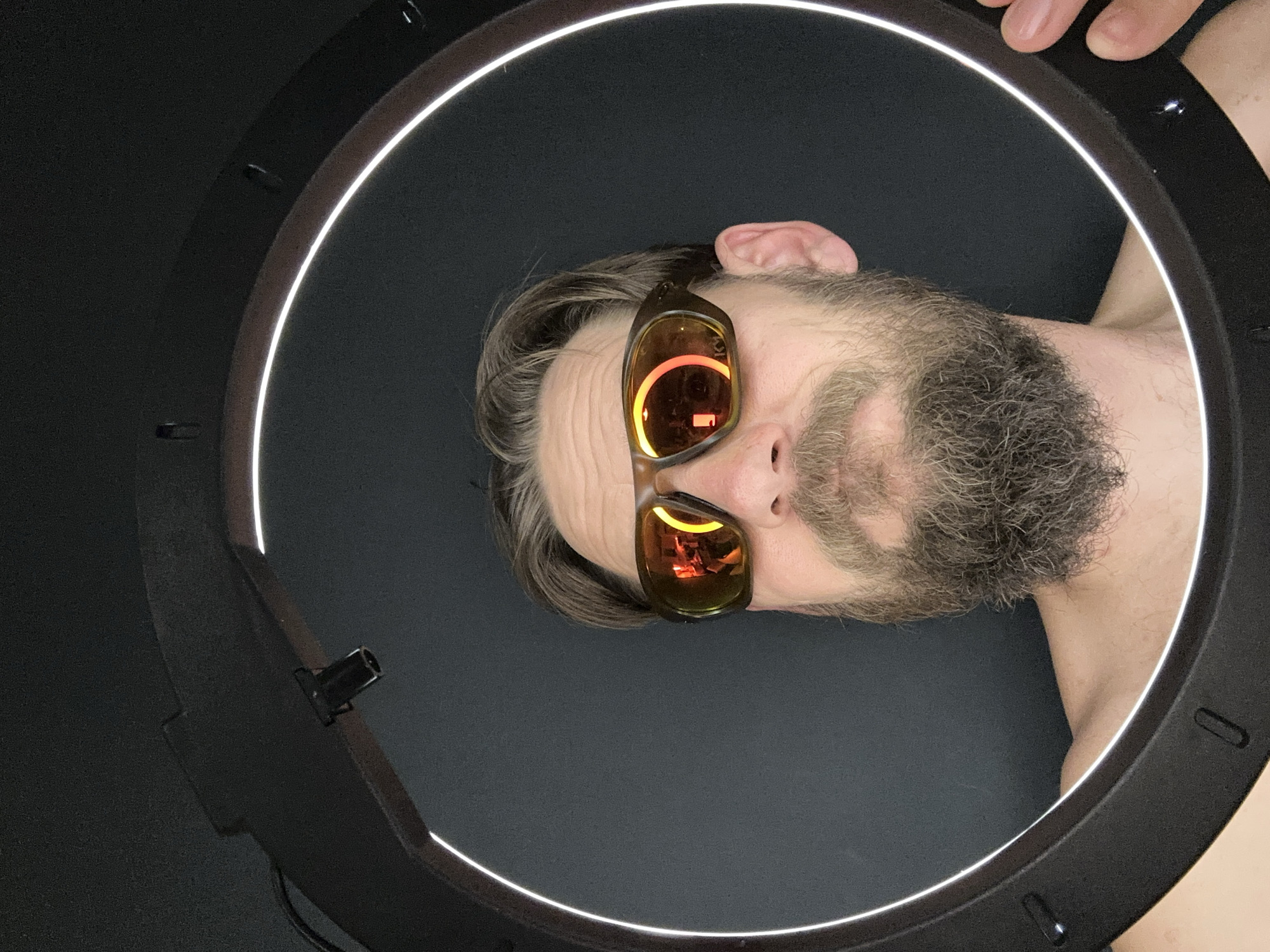A man with a beard, sunglasses, and graying hair appears to be in the center of a ring light. The scene is indoors with a black background. The ring light is black and white, casting light onto the subject's face and creating reflections in the sunglasses. The location in Fethiye, Turkey, is not evident from the image.