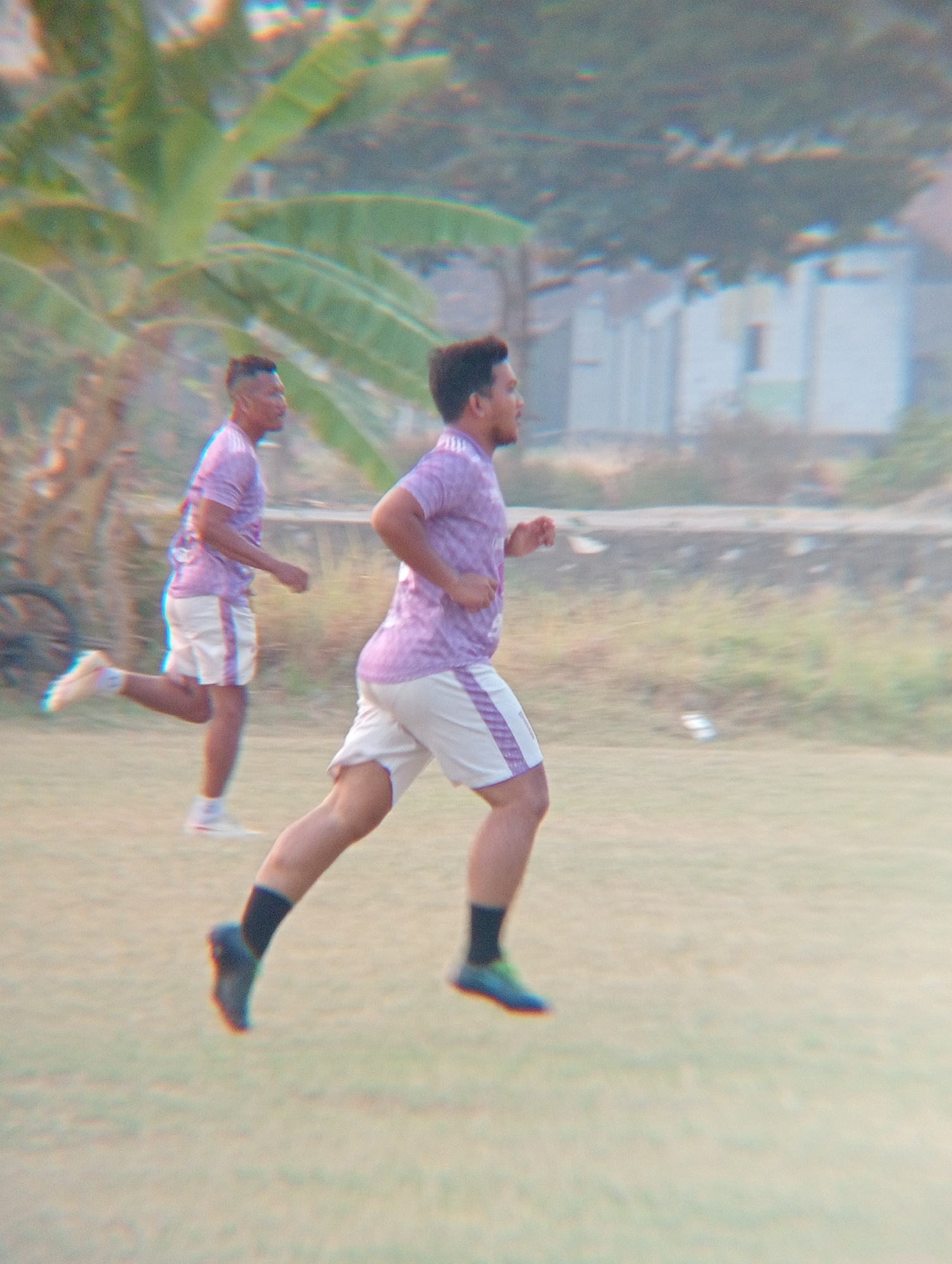 Two men are running on a grassy field in what appears to be a park or outdoor sports facility. The man in the foreground is wearing a purple t-shirt with a patterned design, white shorts with purple stripes, black socks, and blue and green athletic shoes. The man in the background is wearing a similar purple t-shirt and white shorts. Both men are captured in motion, suggesting they are engaged in a race or training exercise. The environment is outdoors, with a blurred background that includes foliage, trees, and some buildings. The lighting suggests it is daytime, possibly late afternoon or early morning, given the soft light. The image has a somewhat hazy or filtered appearance, possibly due to atmospheric conditions or post-processing. There is a bicycle wheel visible in the far left of the background, and the overall impression is one of casual athletic activity in a semi-urban or rural setting. It is not possible to definitively identify the location as Kenep, Indonesia, based solely on the visual information provided.