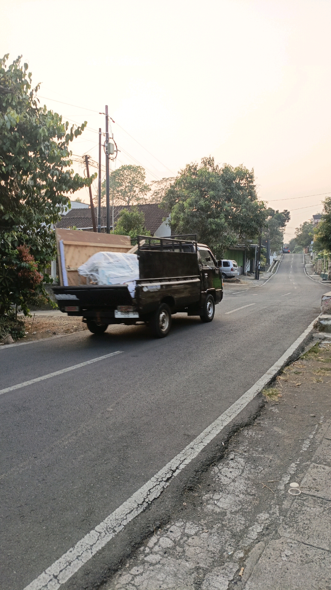 A black pickup truck is driving down a paved road. The truck is carrying large wooden planks and a white tarp-covered object in its bed. The truck is on the left side of the road, and the road curves to the right and uphill. Trees and houses are visible on either side of the road. The sky is a pale, hazy orange, suggesting either dawn or dusk. The scene appears to be in a rural or suburban area. This image was taken in the area of Jalan Gatot Subroto, Sidomulyo, Sidorejo, Salatiga, Indonesia.