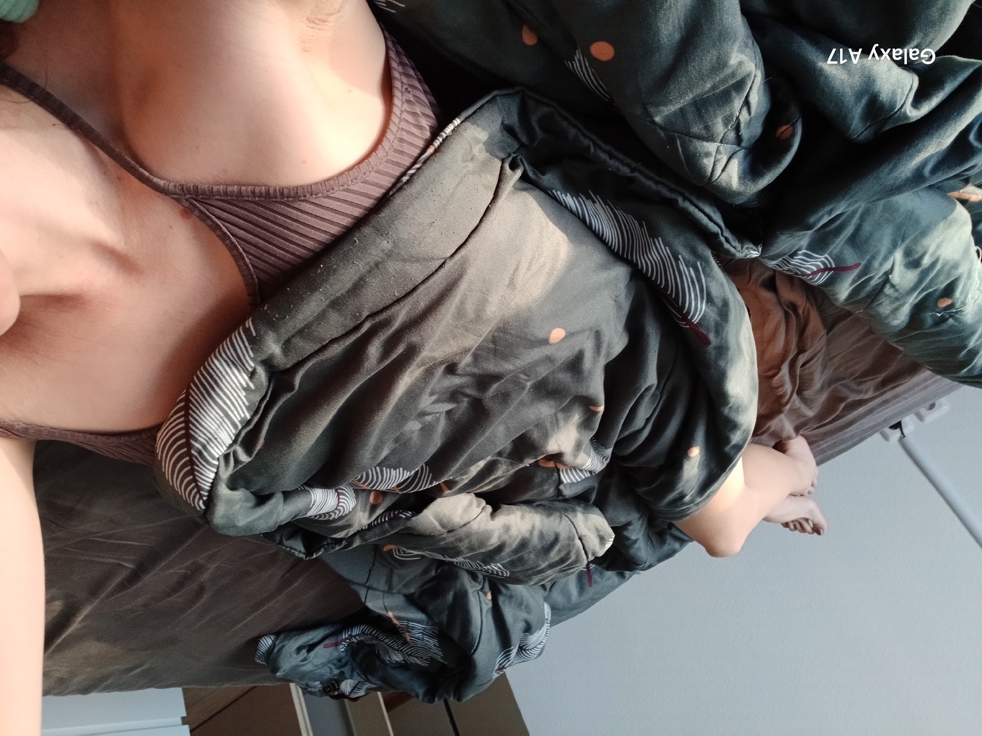 The image shows a person lying in bed under a dark grey comforter with a white and brown patterned design. The person is wearing a brown tank top. The bed has a brown fitted sheet. Part of the person's legs and feet are visible extending out from under the comforter. The background appears to be a bedroom with a white wall and part of a white closet or shelving unit. The text "Galaxy A17" is digitally stamped in the upper right corner. The image was taken in an indoor setting, likely a private residence. There is no information in the image to confirm the location is in Yekaterinburg, Russia.