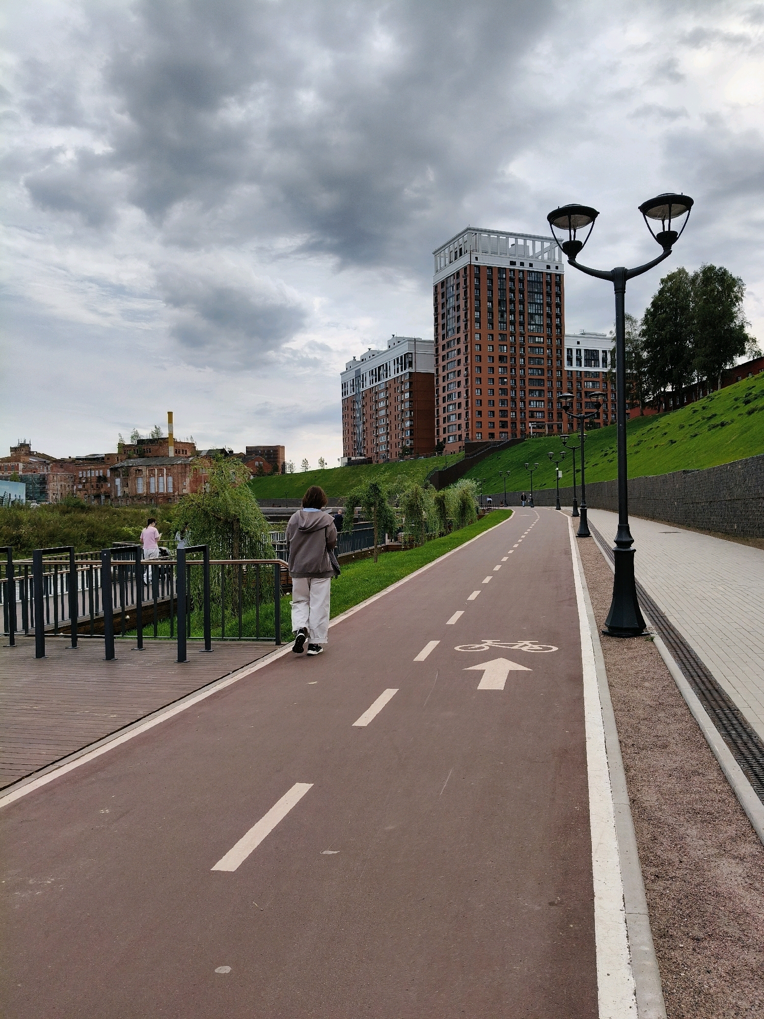 The image depicts an outdoor scene with a paved pathway, likely a bike path or walkway, in Naro-Fominsk, Russia. The path is a reddish-brown color with white dashed lines and a white arrow with a bicycle symbol painted on it, indicating a designated bike lane. A person, viewed from behind, walks away from the camera on this path, dressed in a grey hooded jacket and light-colored pants. To the left of the path is a wooden walkway with metal railings, and beyond that, lush green vegetation. In the background, several tall, modern apartment buildings with red brick facades and white trim are visible. Further in the distance, on the left side of the image, there are older, industrial-looking brick buildings with yellow accents. A grassy hill with some trees rises to the right of the apartment buildings. The sky is overcast with heavy grey clouds, suggesting a cloudy day. Several black lampposts, characteristic of street lighting, are spaced along the right side of the pathway.