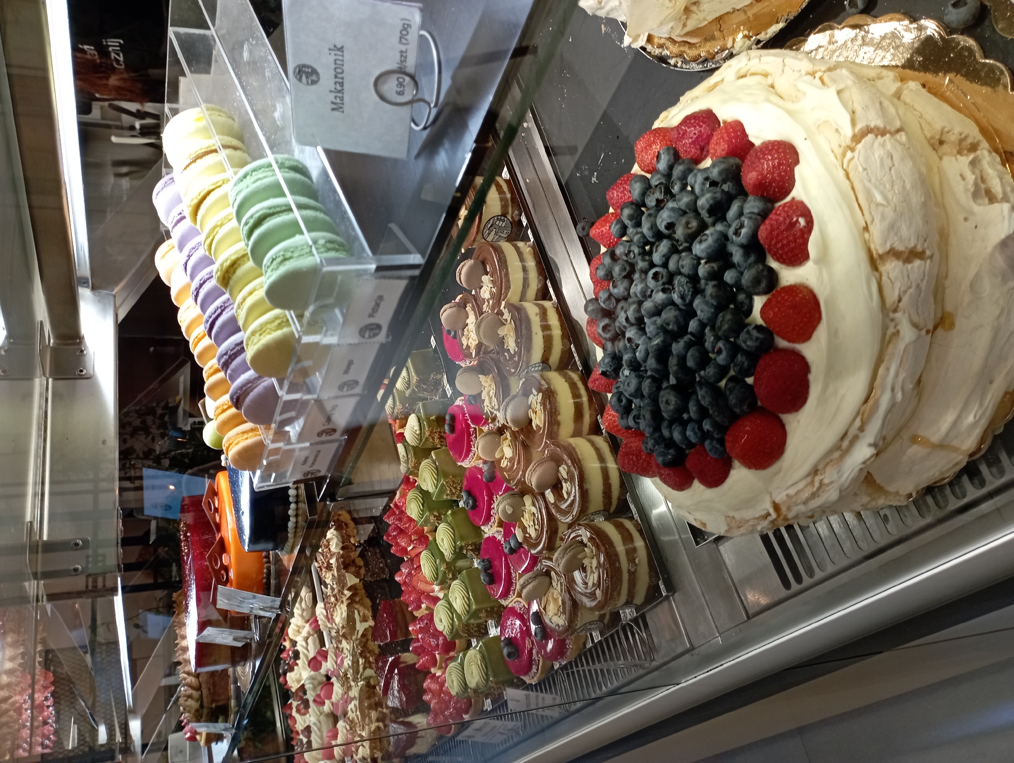 The image displays a refrigerated display case filled with an assortment of pastries, likely at a bakery or patisserie in Katowice, Poland. In the foreground, a large pavlova-style cake is prominently featured, topped with a generous amount of blueberries and strawberries. Behind it, various individual tarts and cakes are arranged, including chocolate layered cakes, fruit-topped desserts, and small green mousse cakes. Further back, rows of colorful macarons are visible in plastic containers, labeled as "Makaronik". The display case is made of glass and illuminated by overhead lights, creating reflections on the surfaces. The background shows shelves of more baked goods and a glimpse of what appears to be a store interior. The overall scene suggests a retail environment focused on selling sweets.