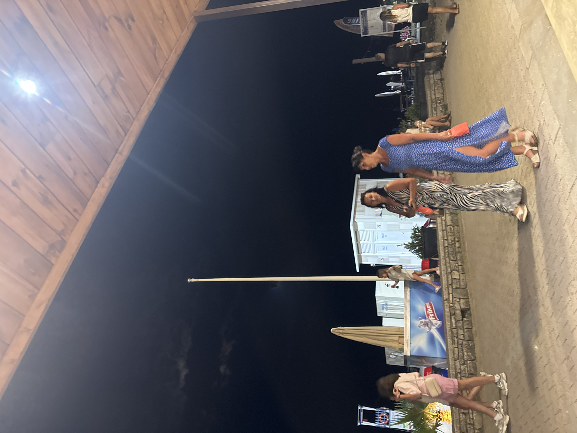 This image, taken at night in Boreti, Montenegro, depicts several people walking along a paved walkway under a wooden overhang. The scene is outdoors and appears to be a commercial area, possibly a promenade or marketplace, given the presence of buildings with what appear to be public restrooms labeled "TOILET".
In the foreground, a woman in a blue, patterned dress and sandals is walking. Behind her, another woman in a black and white zebra-print dress is also walking. Further back, a group of people are visible in the distance, some walking and others standing. One person is wearing a light-colored top and shorts, while another is in dark clothing. A child in a light pink top and pink shorts is also present, walking with a light-colored bag over their shoulder.
The time of day is clearly night, indicated by the dark sky and artificial lighting. The weather appears to be clear, with no visible signs of rain or wind. The presence of umbrellas suggests a place that caters to outdoor activities, perhaps restaurants or beachside services.
A prominent sign in the background displays the word "Frikom", likely a brand name, and an image of a polar bear. The overall mood of the image seems casual and relaxed, with people enjoying an evening stroll. The wooden structure in the foreground suggests a covered area, possibly part of a building or an awning, offering shade or shelter.