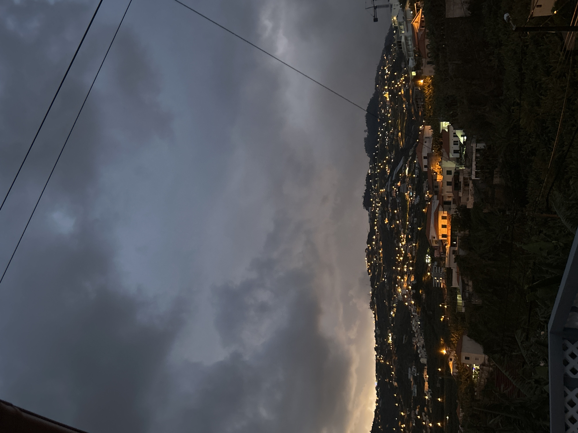 The image depicts a hillside townscape in Câmara de Lobos, Portugal, at twilight. The sky is overcast with dark, heavy clouds. Scattered lights illuminate the houses and buildings on the steep hillside. A few electrical lines cross the top part of the frame. Lush vegetation flanks the buildings. A section of a railing appears in the foreground.