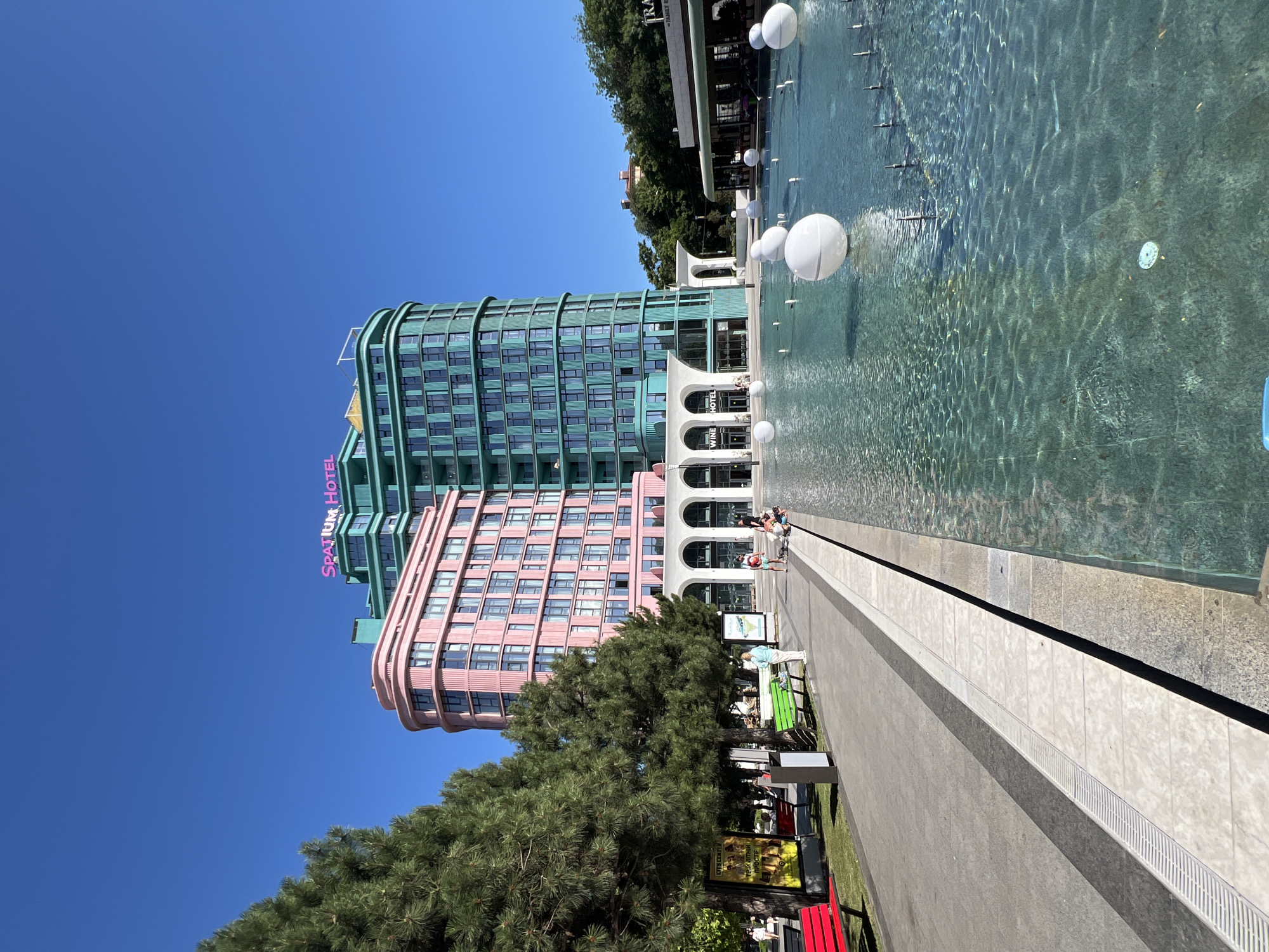 The image captures a sunny day in Odesa, Ukraine, in front of the Spatium Hotel. The hotel's architecture is distinctive, with one section painted pink and another teal, both featuring modern design elements and numerous windows. A bright pink sign on top of the hotel clearly reads "SPATIUM HOTEL".
In the foreground, a large, shallow fountain dominates the scene. Several large white spheres float on the water's surface, and smaller water jets are visible. The water itself is clear and reflects the bright blue sky. A paved walkway runs alongside the fountain, with a row of trees to the left of the walkway, providing shade. Several benches, some painted red and green, are placed along the path.
A few people are scattered throughout the scene. Some are walking along the path, while others are seated or interacting near the fountain. On the right side of the image, a building with a sign that appears to say "TRA..." and possibly "FAMILY" is visible, suggesting it might be a cafe or restaurant. The overall atmosphere is one of a pleasant urban public space on a warm day.