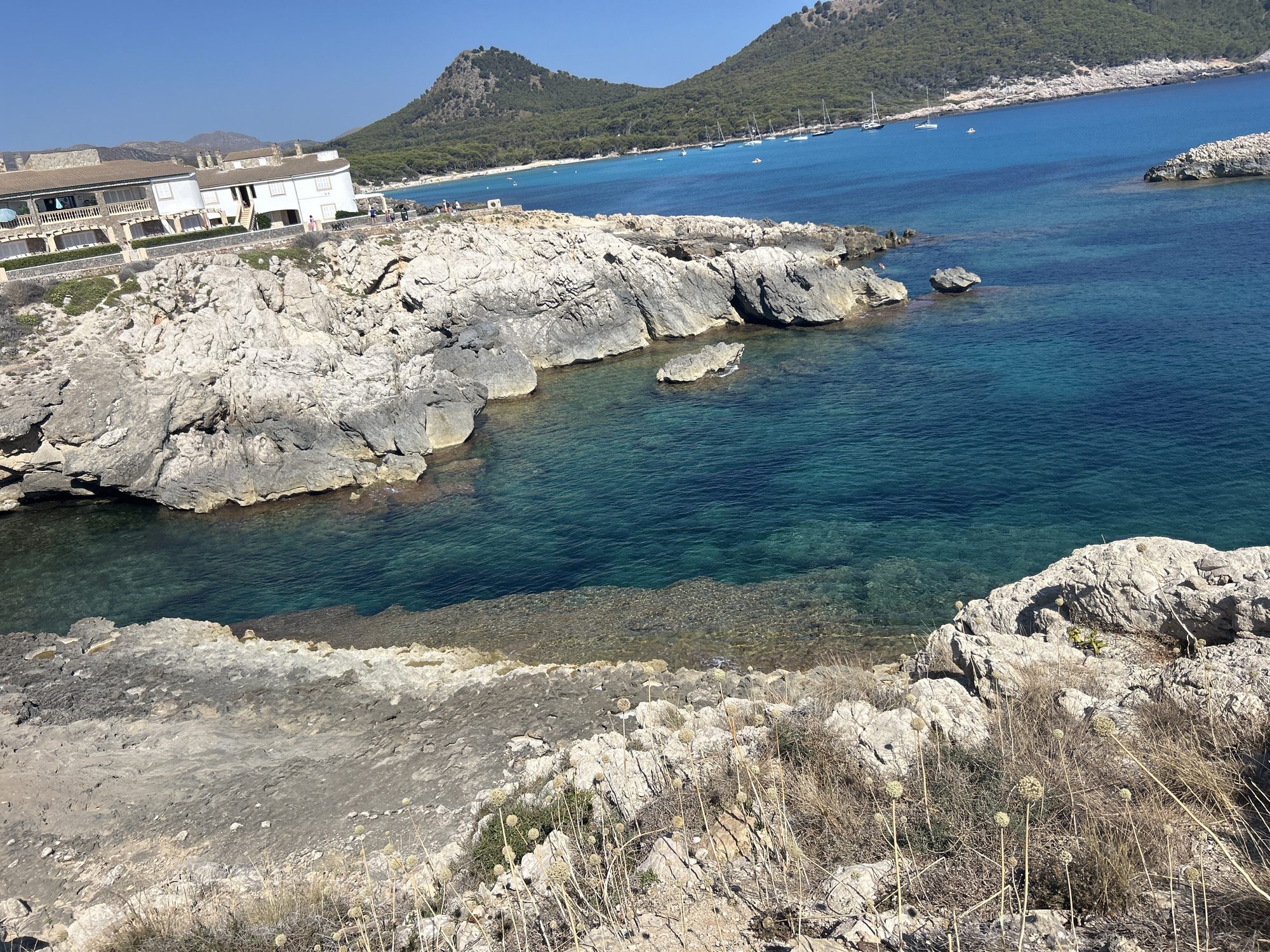 This is a high-angle, eye-level, wide shot photograph of a coastal scene in Cala Lliteres, Spain. The foreground is dominated by a rugged, rocky shoreline with dry, sparse vegetation. Beyond this, a calm, clear turquoise sea stretches out. In the midground, a bay is visible with several sailboats anchored. Along the coastline of the bay, there is a white building complex with multiple levels and balconies, situated on a terraced hillside. Further in the distance, lush green hills rise, and the sky is a clear, bright blue. There are several people visible on a pathway near the white buildings, and a few individuals are swimming or wading in the sea near the rocky outcrops. The overall impression is of a serene and picturesque Mediterranean landscape.