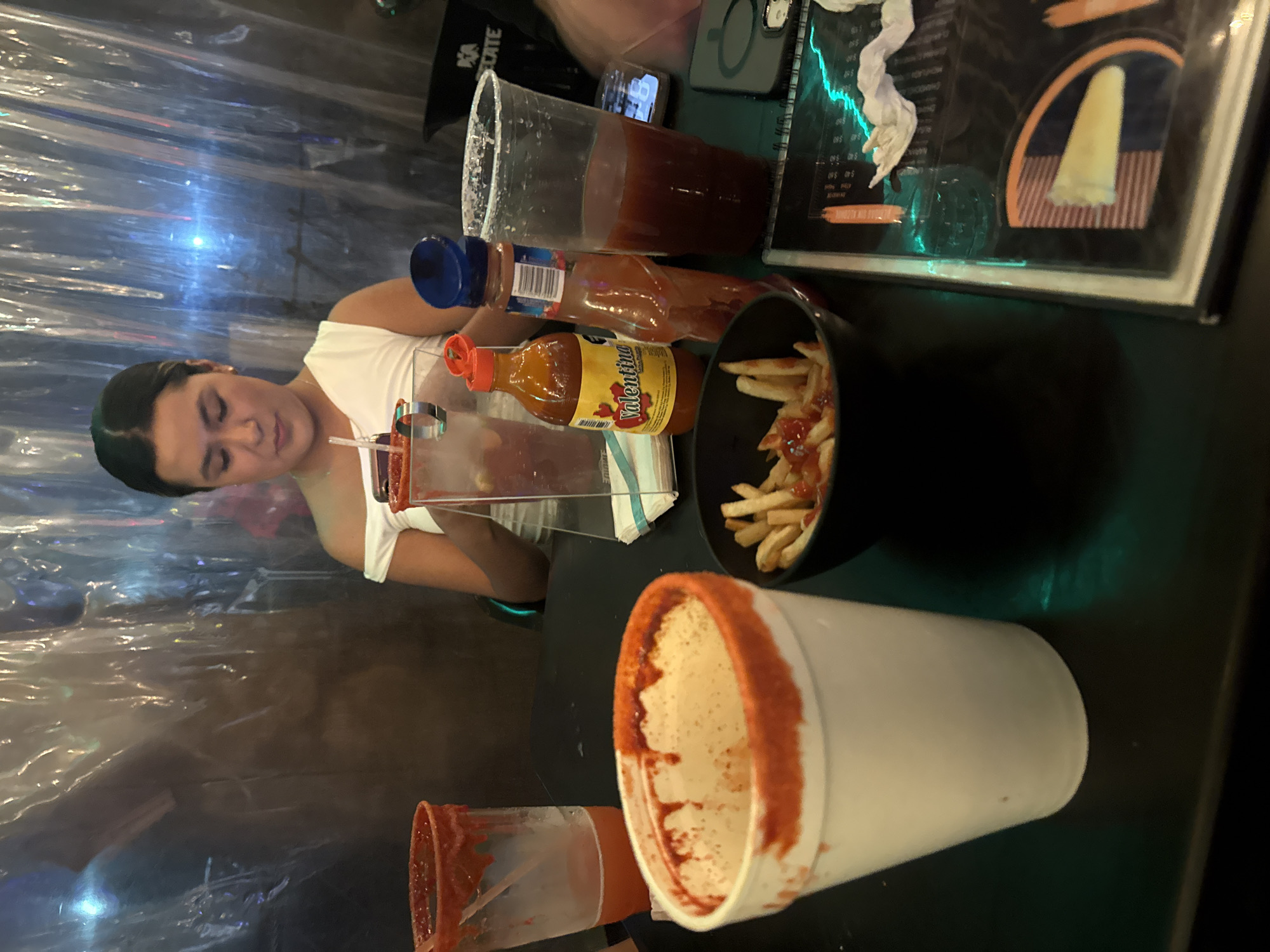 A woman is sitting at a table in what appears to be a restaurant or bar. The location is indicated as San Bernardino Tlaxcalancingo, Mexico, though this is based on external information and not discernible from the image itself.
The table contains several items:
* A tall, clear plastic cup filled with a red liquid, likely a beverage.
* Two bottles of Valentina hot sauce.
* A small bowl of French fries, some with ketchup.
* Another rimmed cup.
* A menu.
* A cell phone.
The environment is partially enclosed by clear plastic barriers, likely to separate seating areas.