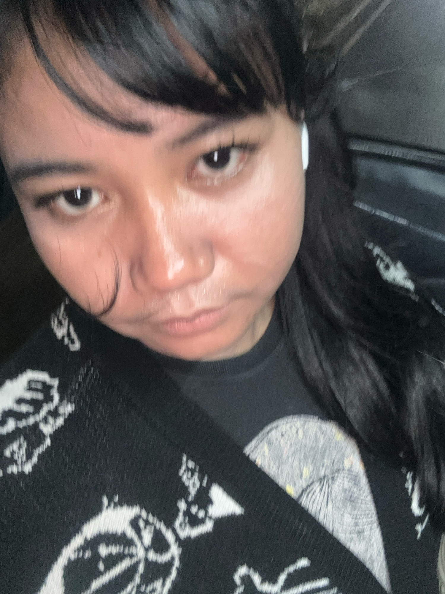 The image shows a close-up, slightly blurred, selfie of a person with long, dark hair and bangs. They are wearing a black t-shirt with a white graphic design and a black cardigan also with white graphic designs. A white earbud is visible in their right ear. The lighting suggests it is indoors, possibly in a vehicle given the dark background with what appears to be textured upholstery. The person's facial expression is neutral to slightly pensive. There are no discernible landmarks or features that would indicate a specific location within North Jakarta, Indonesia.