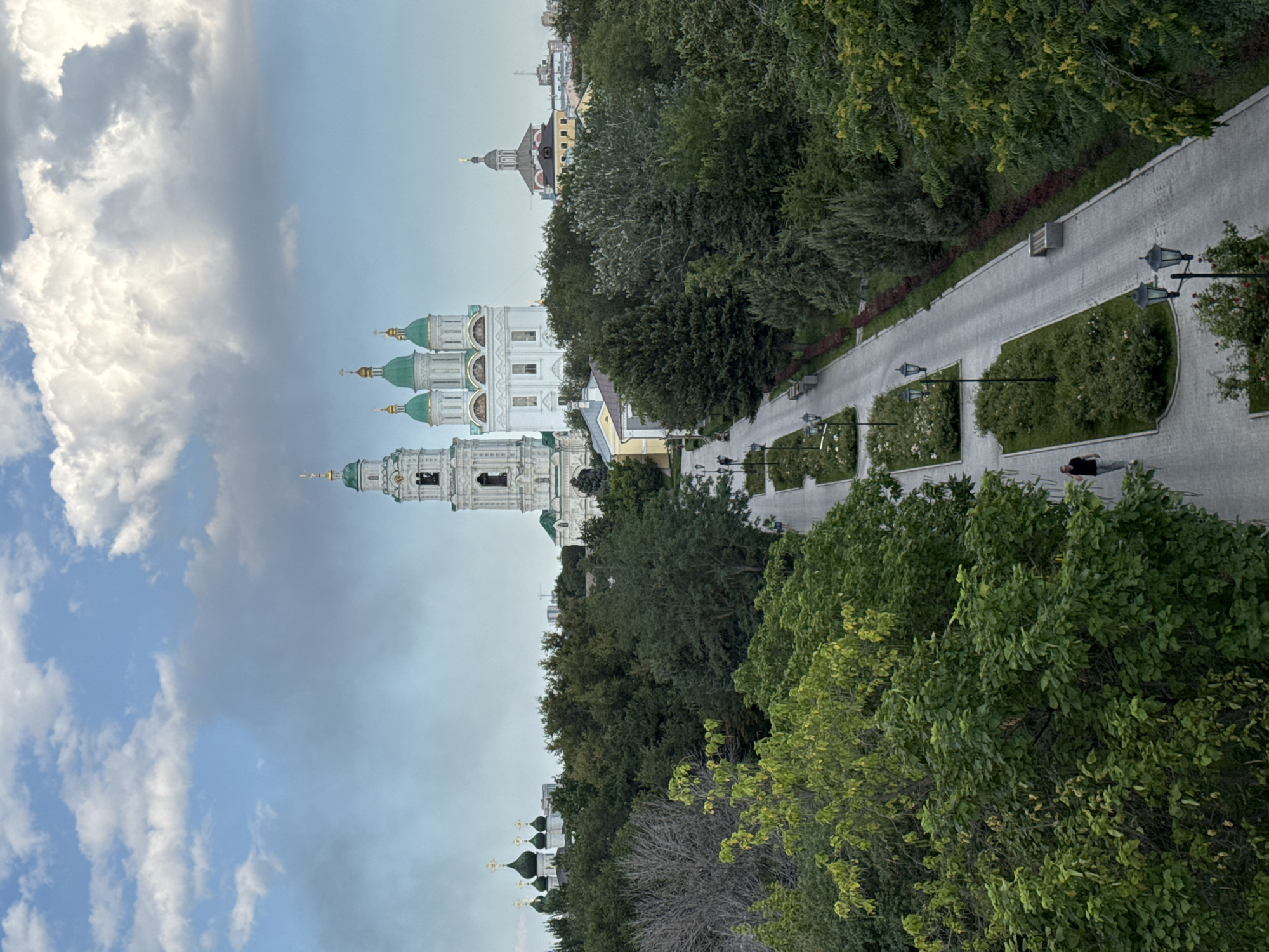The image depicts a view looking down a tree-lined pathway in Astrakhan, Russia, leading towards the Assumption Cathedral and bell tower, iconic landmarks of the Astrakhan Kremlin. The pathway, paved with grey material, is flanked by meticulously maintained green lawns with decorative flowerbeds. Several lampposts are positioned along the path, and benches are visible on the right side. A lone individual walks on the pathway in the middle distance. Beyond the pathway and a dense green forest, the white and green domed Assumption Cathedral and its tall bell tower rise majestically. Several other smaller, green-domed religious structures and buildings are visible in the background, interspersed with trees and the urban landscape of Astrakhan. The sky above is a mix of blue with large, fluffy white clouds. The overall scene suggests a pleasant day in a well-preserved historical and religious complex.