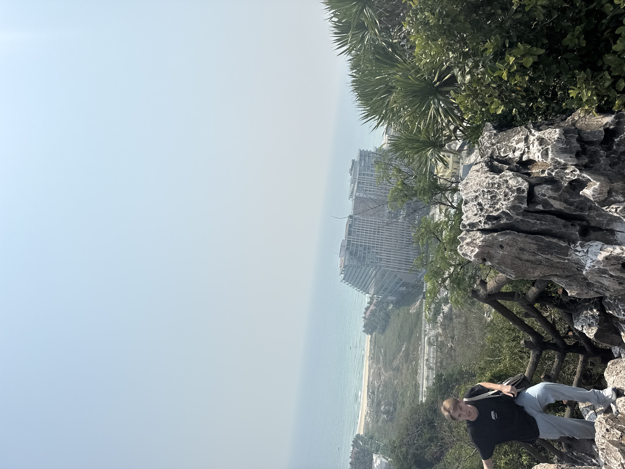 A person stands on a rocky outcrop along a railing. A multi-story building stands in the background, near the water. The sky is hazy. The scene may be in Đà Nẵng, Vietnam.