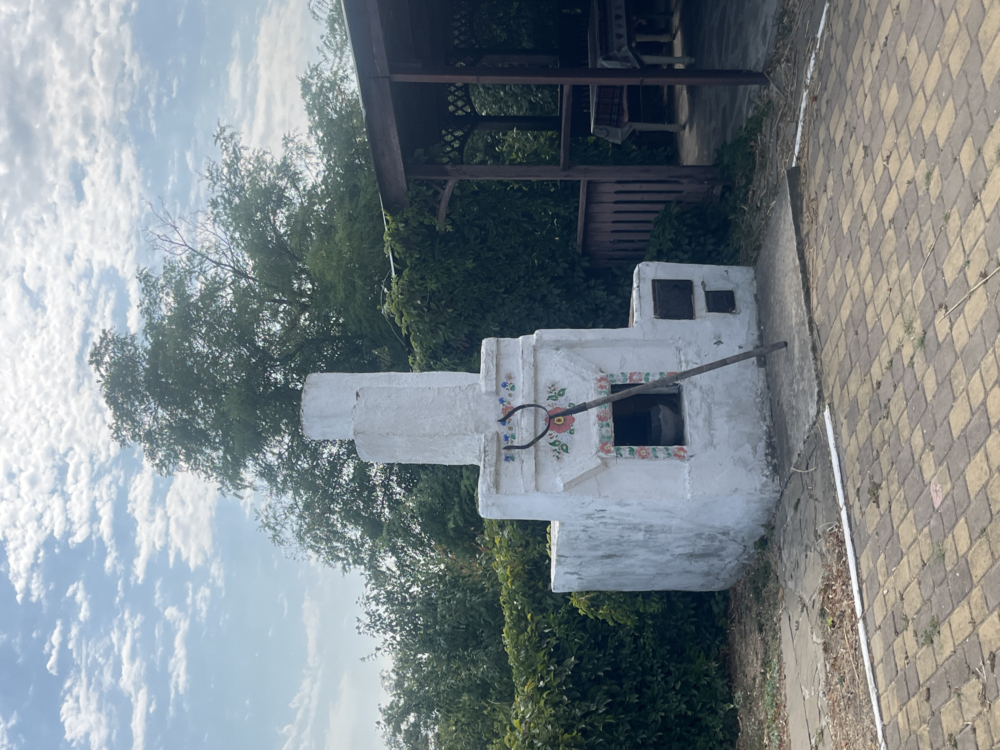 A white, outdoor oven with colorful floral decorations stands on a paved patio in Taman', Russia. The oven is tall and rectangular, with a chimney. A long, dark tool, possibly a poker or shovel, rests against the oven. Behind the oven, a wooden gazebo with a table and chairs is visible. Lush green bushes and trees surround the area, and a cloudy sky can be seen above. The ground is a mixture of paving stones and dry earth.