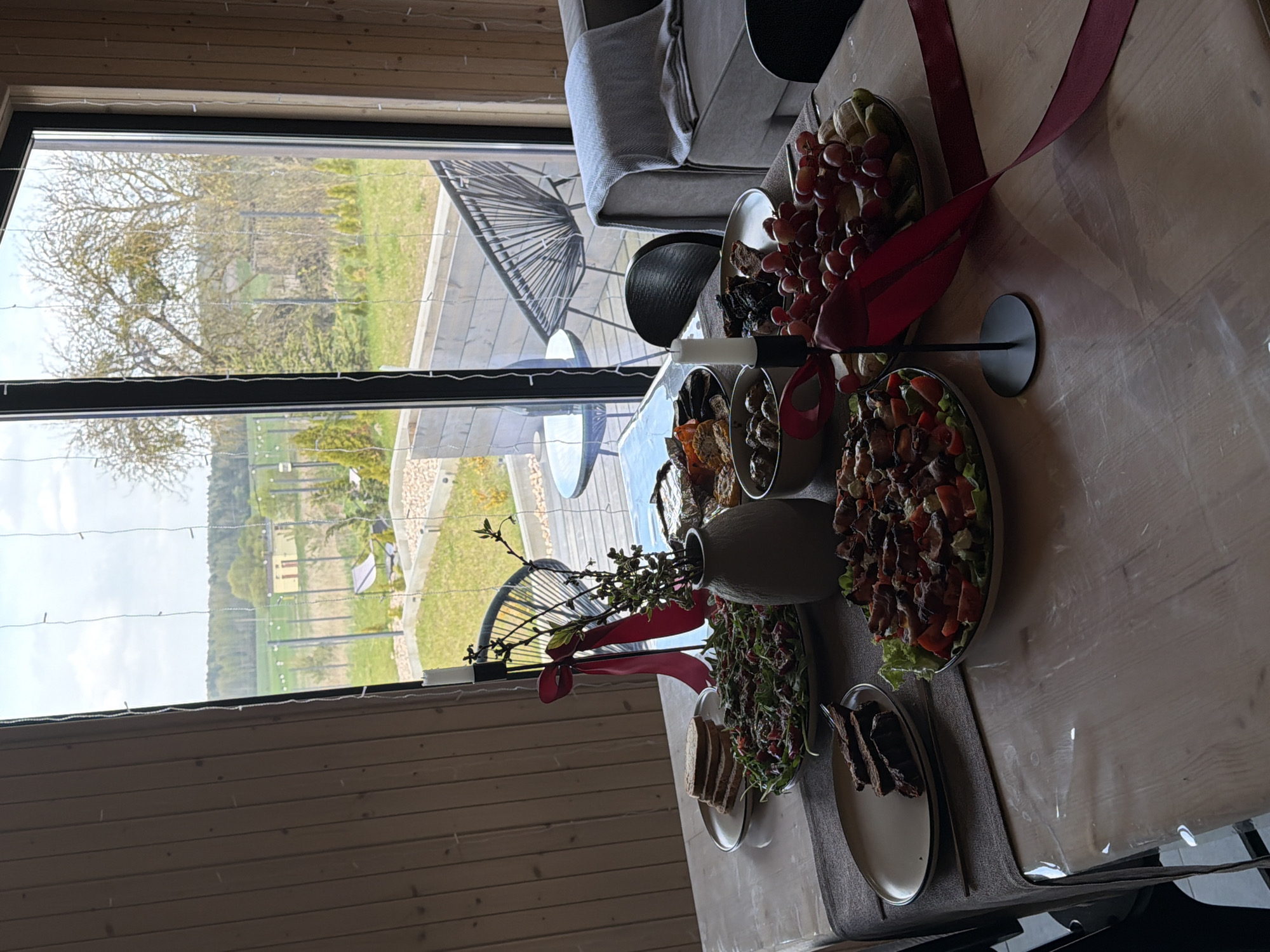 The image shows a table laden with food inside a room, with a large window providing a view of the exterior.
**Location:** The scene is set indoors, within a room that appears to be part of a house or similar dwelling, based on the window view and interior design. The location is specified as Капцёўскі сельскі Савет, Belarus.
**Objects:**
* A table, covered with food in serving dishes and plates.
* Bowls and plates containing grapes, meat, olives, salads, and slices of bread.
* A vase holding greenery and tied with a red ribbon.
* A sofa visible near the window.
* Chairs next to the table.
* A large window with a view of the outside.
**Environment:**
* The room has wooden walls.
* Outside, a landscape with grass, trees, and fields can be seen through the window.
* The weather appears to be overcast or cloudy.
**Subject:**
* Food arrangement on a table indoors.
**Actions:**
* Food is arranged on the table in preparation for a meal or gathering.
**Interactions:**
* The image suggests a social event or meal is about to take place, as the table is set with a variety of food items.