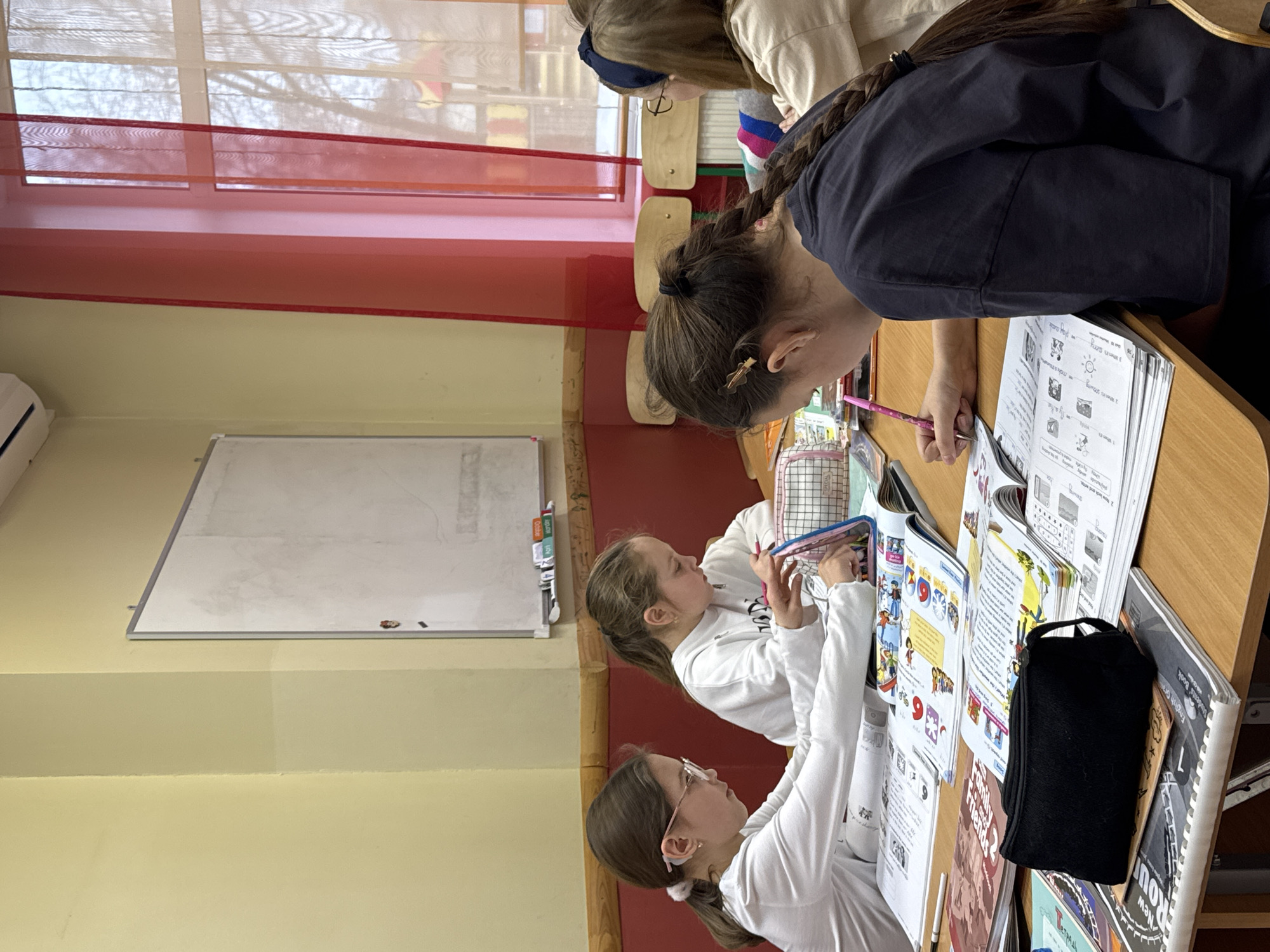 The image captures a scene inside a classroom, likely in Perm, Russia (based on the notebook labeled "Tetrad'" which uses the Russian Cyrillic alphabet). Several young students are seated at desks, apparently engaged in their schoolwork. One girl is leaning over her desk, writing in a textbook with a pink pencil. In front of her are several open textbooks. A younger girl in front of her is also occupied with school materials. The classroom features a whiteboard on the wall, a light yellow wall color, and windows with red curtains. Other students are visible in the background. The desks are wooden, and various school supplies are scattered around.