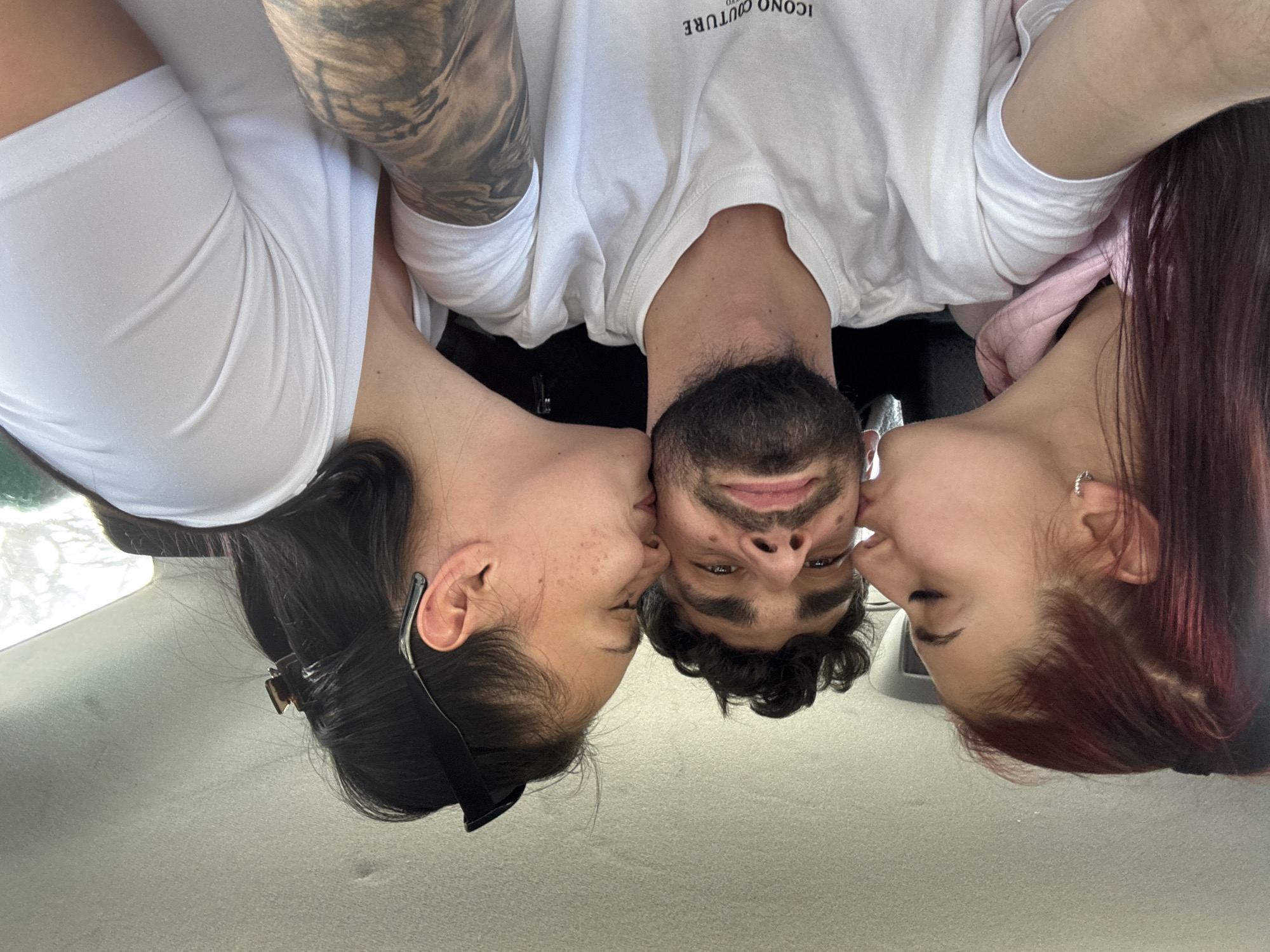 A low-angle, upside-down photograph depicts a man being kissed on each cheek by two women. The man has dark hair, a beard, and is wearing a white t-shirt. The woman on the left has dark hair pulled back with a hair clip and is wearing a white top. The woman on the right has reddish hair and is wearing a pink shirt. The location appears to be inside a vehicle, as suggested by the car's interior. The city and specific location are unknown.