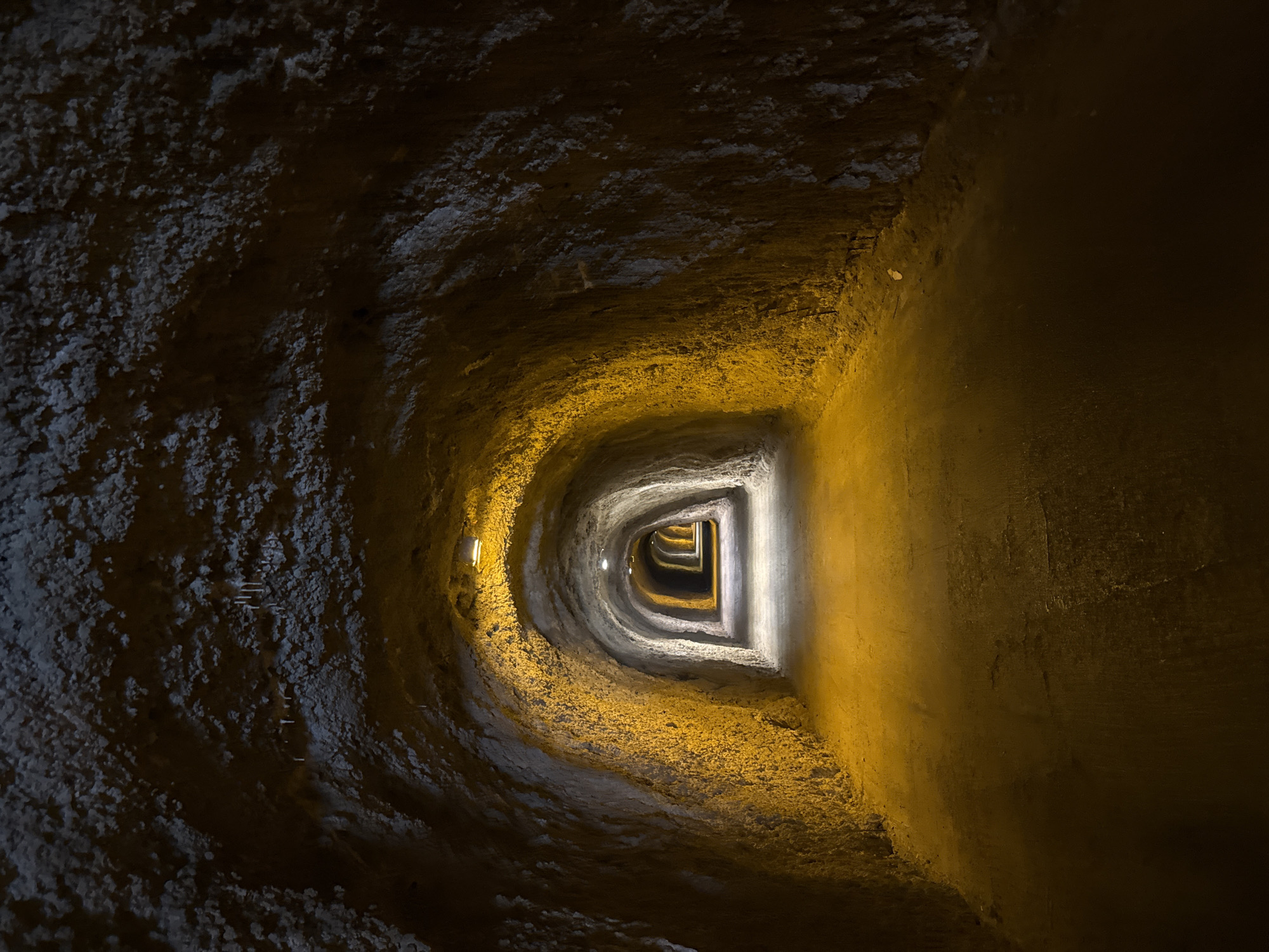The image depicts the interior of a tunnel with rough, uneven walls that are illuminated by artificial lighting. The tunnel appears to be carved out of rock or earth, with textures suggesting a natural or crudely excavated formation. Small, elongated formations resembling stalactites hang from the ceiling in some areas. The tunnel recedes into the distance, with multiple visible arches or openings creating a sense of depth and repetition. The lighting is predominantly yellow-orange, casting a warm glow on the surfaces and creating areas of shadow. The floor of the tunnel is a smooth, dark surface, likely a pathway or road. There are no people or animals visible in the scene. The overall impression is one of an underground passage, possibly man-made or a natural cave adapted for passage.
The provided context places this scene in Kamenovo, Montenegro. While the image itself does not contain specific visual cues to confirm this location, it is consistent with the appearance of tunnels that might be found in coastal or mountainous regions, which describe parts of Montenegro.