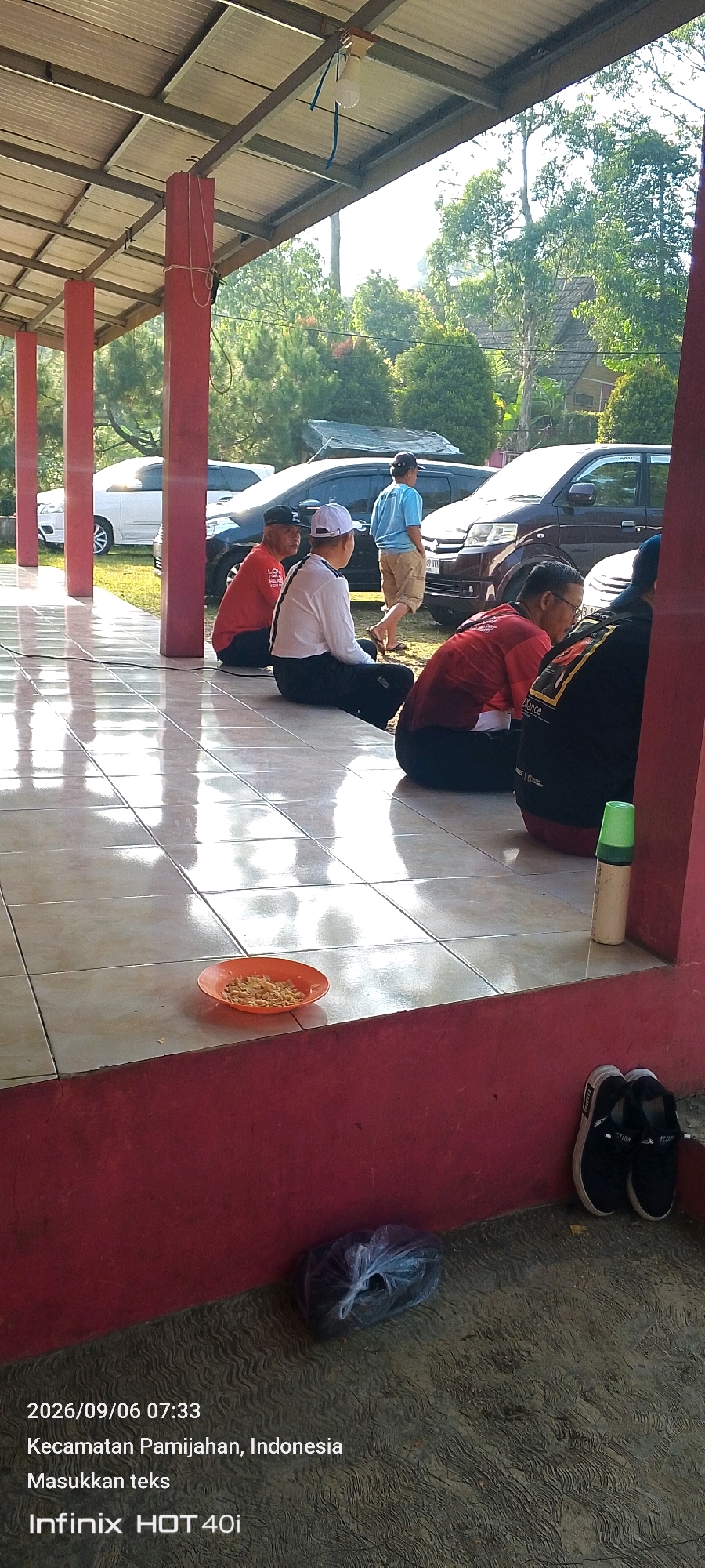 The image depicts an outdoor scene in Pamijahan, Indonesia. Several people are sitting on a tiled floor under a covered patio. The patio has red pillars and a metal roof. In the background, cars are parked, and lush greenery with trees and some buildings are visible. A small orange plate with food is on the tiled floor in the foreground, along with a plastic bag and a pair of black and white sneakers. The timestamp on the image indicates it was taken on September 6, 2026, at 07:33.