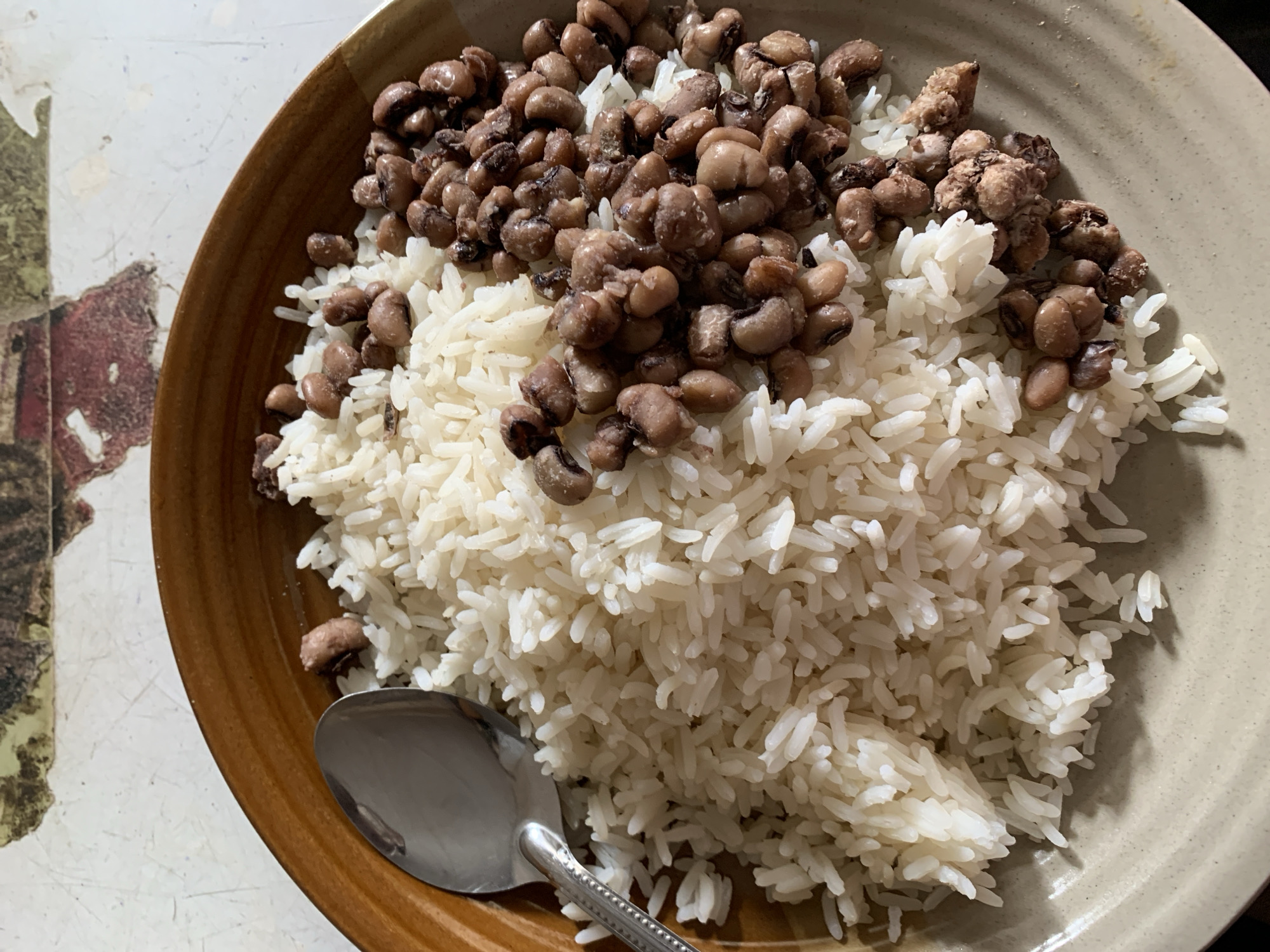 A plate filled with a mound of cooked white rice is topped with cooked black-eyed peas. A silver spoon rests on the edge of the plate. The plate is brown and beige. The setting is unidentified, with a pattern on the table visible near the plate. There is no direct evidence suggesting the location is New Stadium, Nigeria.
