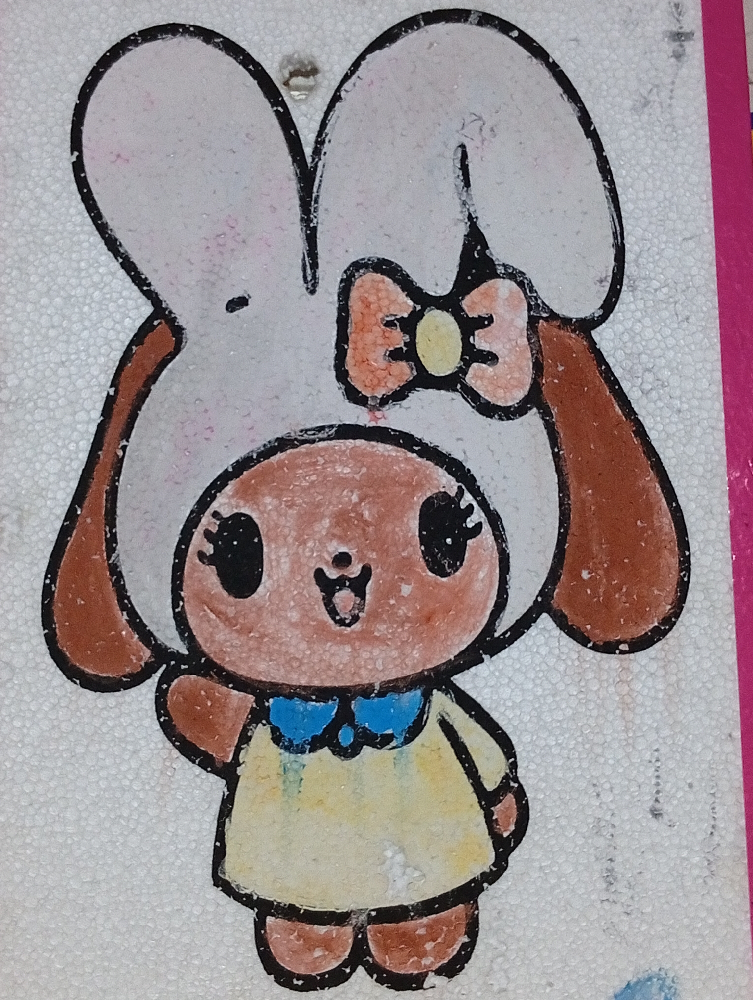This is a photograph of a drawing on a white, textured surface, possibly styrofoam. The drawing depicts a cartoon character resembling a rabbit or a bear wearing a white hood with large ears and a red and orange bow. The character has a pinkish-brown face with black eyes, a small smile, and black eyelashes. It is wearing a yellow dress with a blue collar. The drawing is outlined in black. A bright pink border is visible along the right edge of the image. There is no discernible connection to Lhokseumawe, Indonesia, as the image focuses solely on the artwork itself and provides no contextual environmental or geographical information.