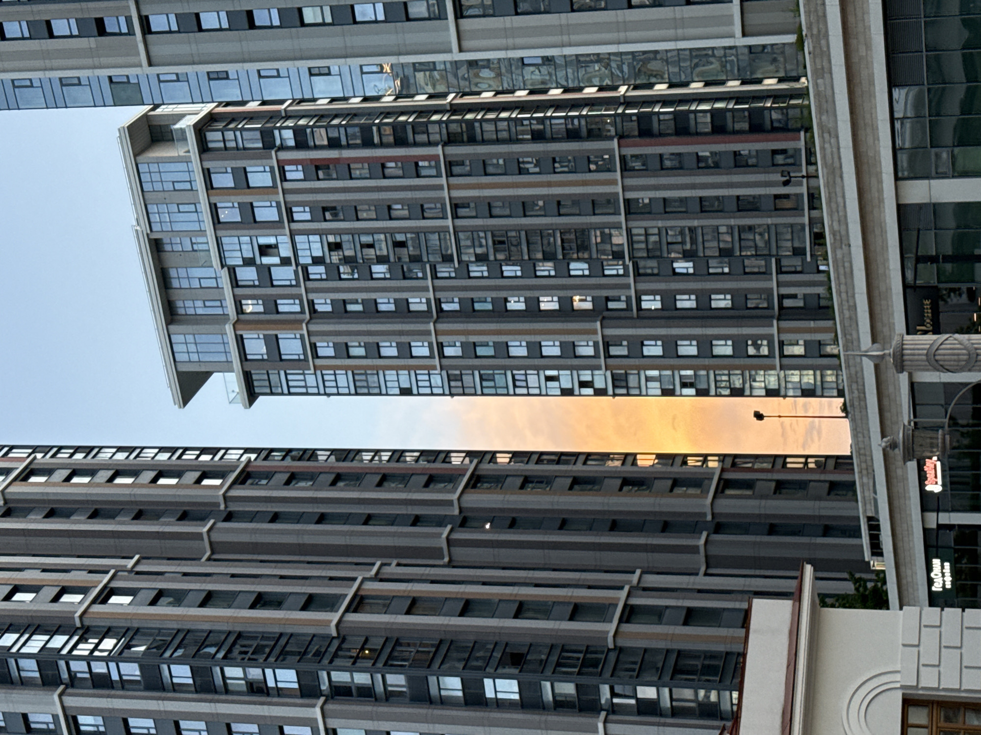The image shows a street view in Moscow, Russia, focusing on a cluster of modern high-rise apartment buildings. The buildings are characterized by their dark, geometric facades with numerous windows, interspersed with vertical bands of lighter gray and occasional thin, colored stripes in shades of orange and red. The sky is a clear, pale blue.
In the foreground, a lower, white building with arched architectural details is visible on the left. Below the apartment buildings, a horizontal structure with large glass windows and signage can be seen. One sign displays Cyrillic text in green, reading "ГенОдиан кофешоп" (GenOdian Coffee Shop), and another, to its right, shows a red logo that appears to be "SberMarket". A lamppost with a single light is positioned between two of the taller buildings, its slender dark pole extending upwards against the orange glow of the sky. The overall impression is one of urban development, with a juxtaposition of older architectural styles in the foreground and contemporary high-rise structures dominating the scene.
